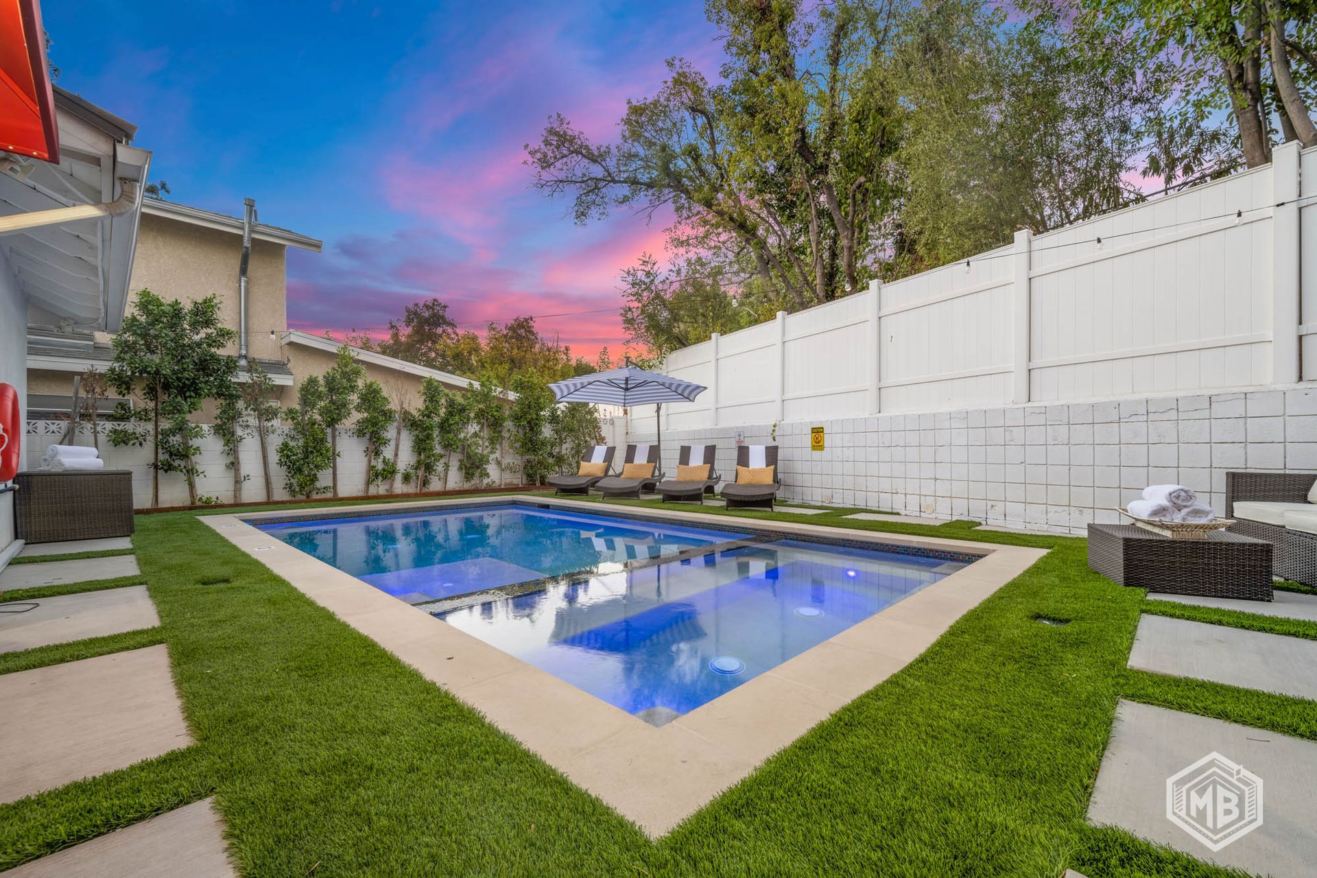 A rectangular backyard swimming pool surrounded by green turf, stone pavers, and a white privacy fence at sunset.