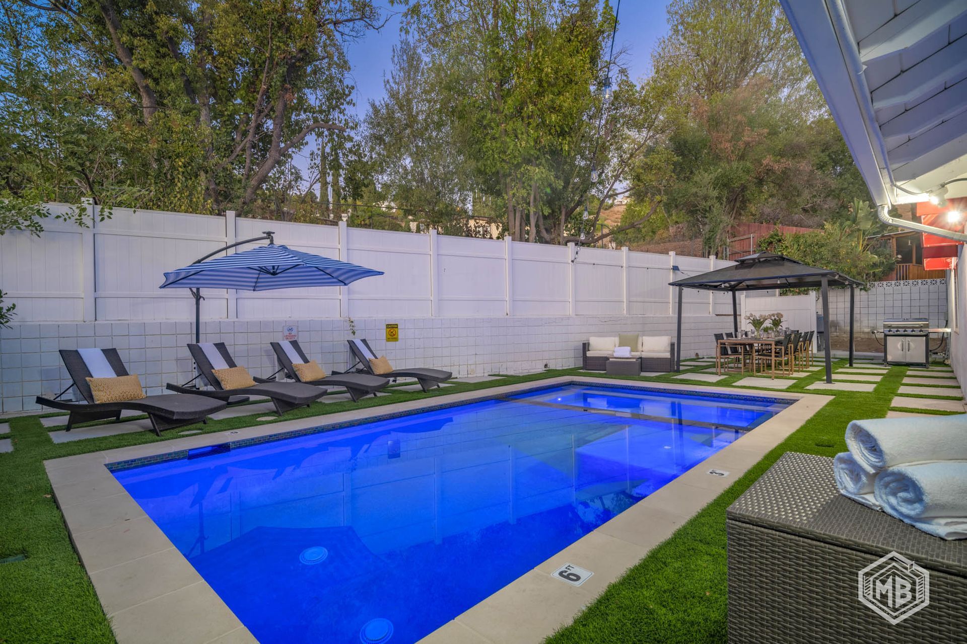 A rectangular backyard swimming pool with blue water surrounded by lounge chairs, an umbrella, and a covered patio area.