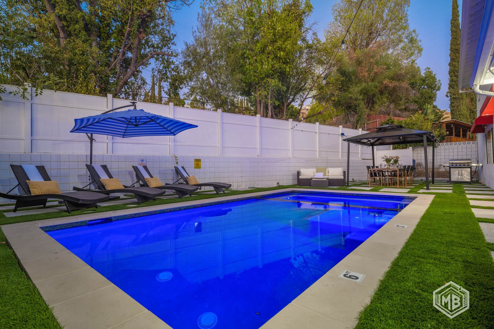 A rectangular swimming pool with blue water sits in a landscaped backyard with lounge chairs and a gazebo.
