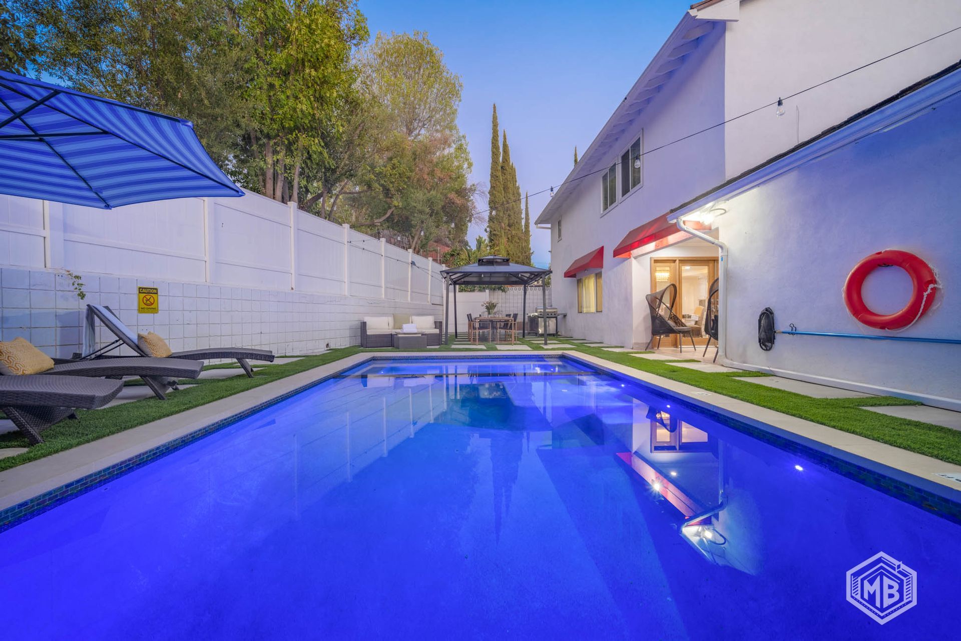 A rectangular backyard swimming pool with blue water, lounge chairs under a striped umbrella, and a house exterior.