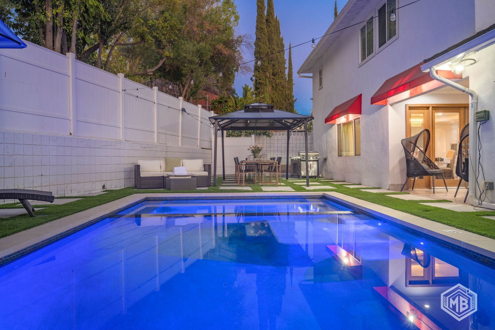 A vibrant blue swimming pool in a backyard at dusk, featuring a patio set, a gazebo, and a white house with red awnings.