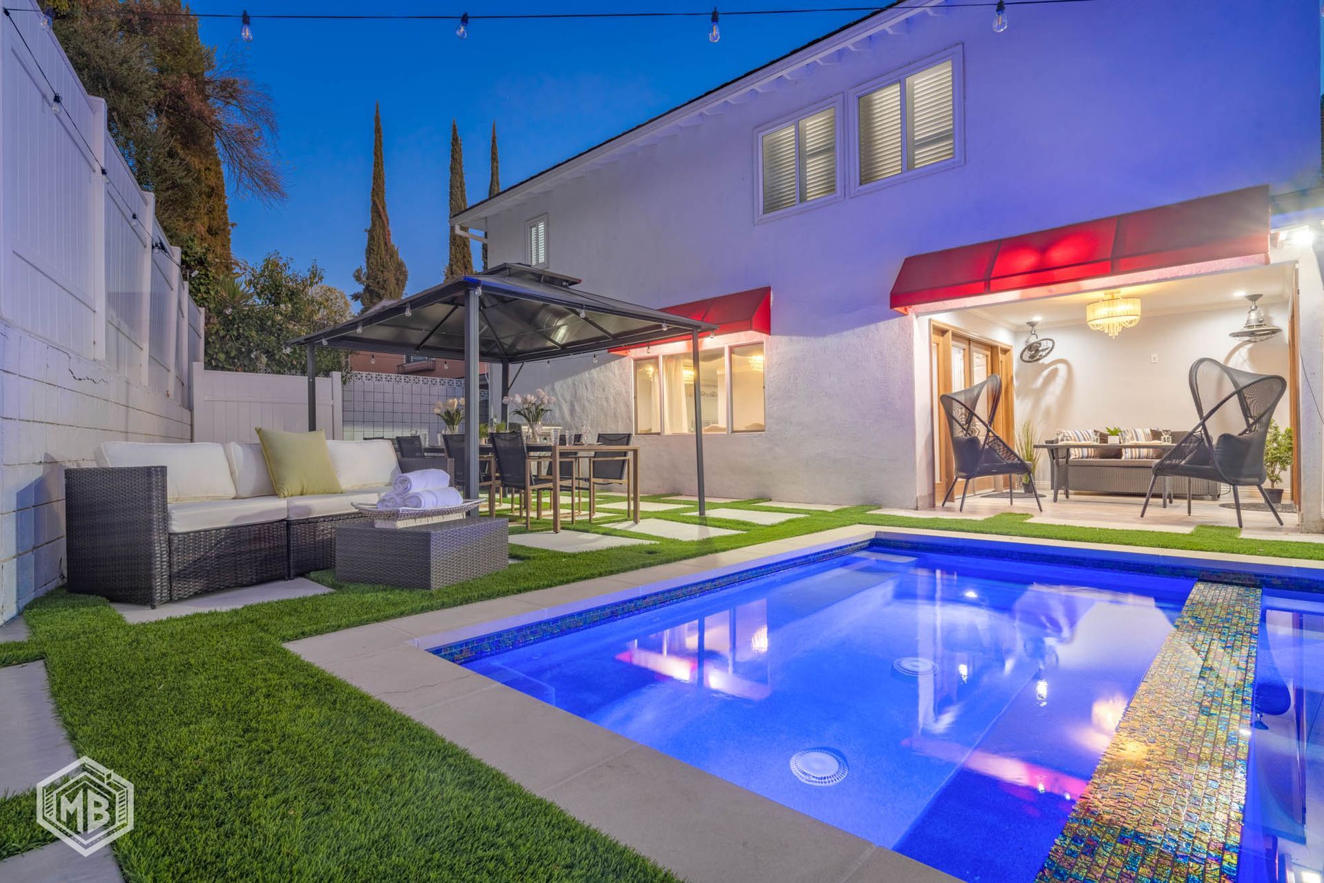 A backyard patio with a rectangular swimming pool, outdoor seating, a gazebo, and a home exterior at dusk.