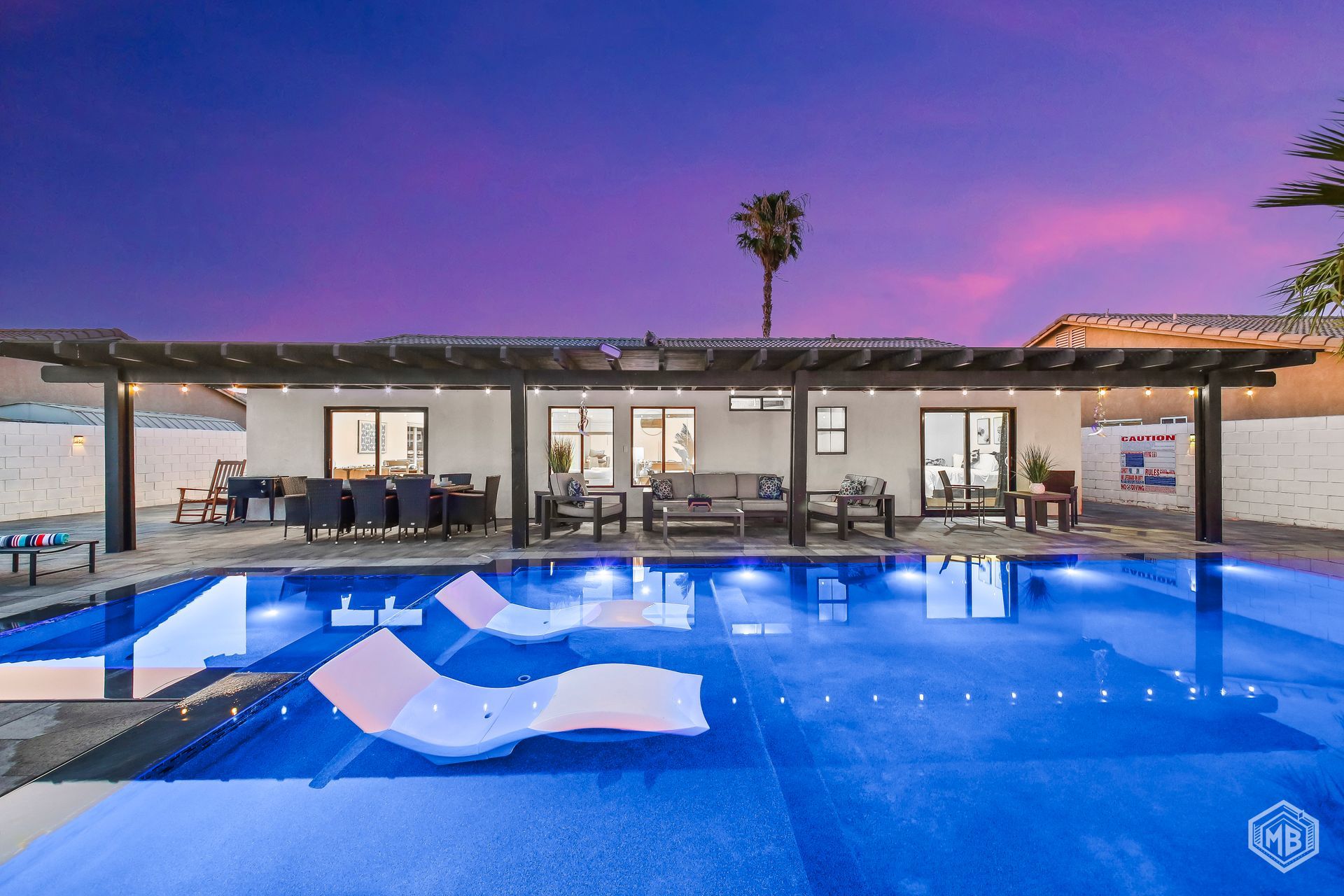 A vibrant twilight sky hangs over a modern patio with lounge chairs and dining space beside a bright blue swimming pool.