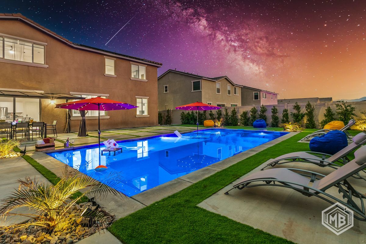 A backyard swimming pool at night, illuminated by blue lights under a vibrant, starry night sky with a milky way view.