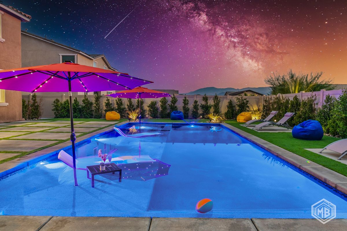 A swimming pool at night under a vibrant, star-filled sky, with a purple umbrella, deck chairs, and a colorful ball.