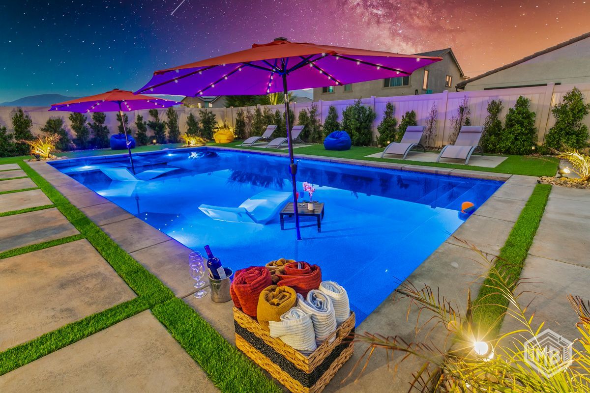 A pool at night, illuminated with blue lights, featuring floating umbrellas, lounge chairs, and a basket of towels.
