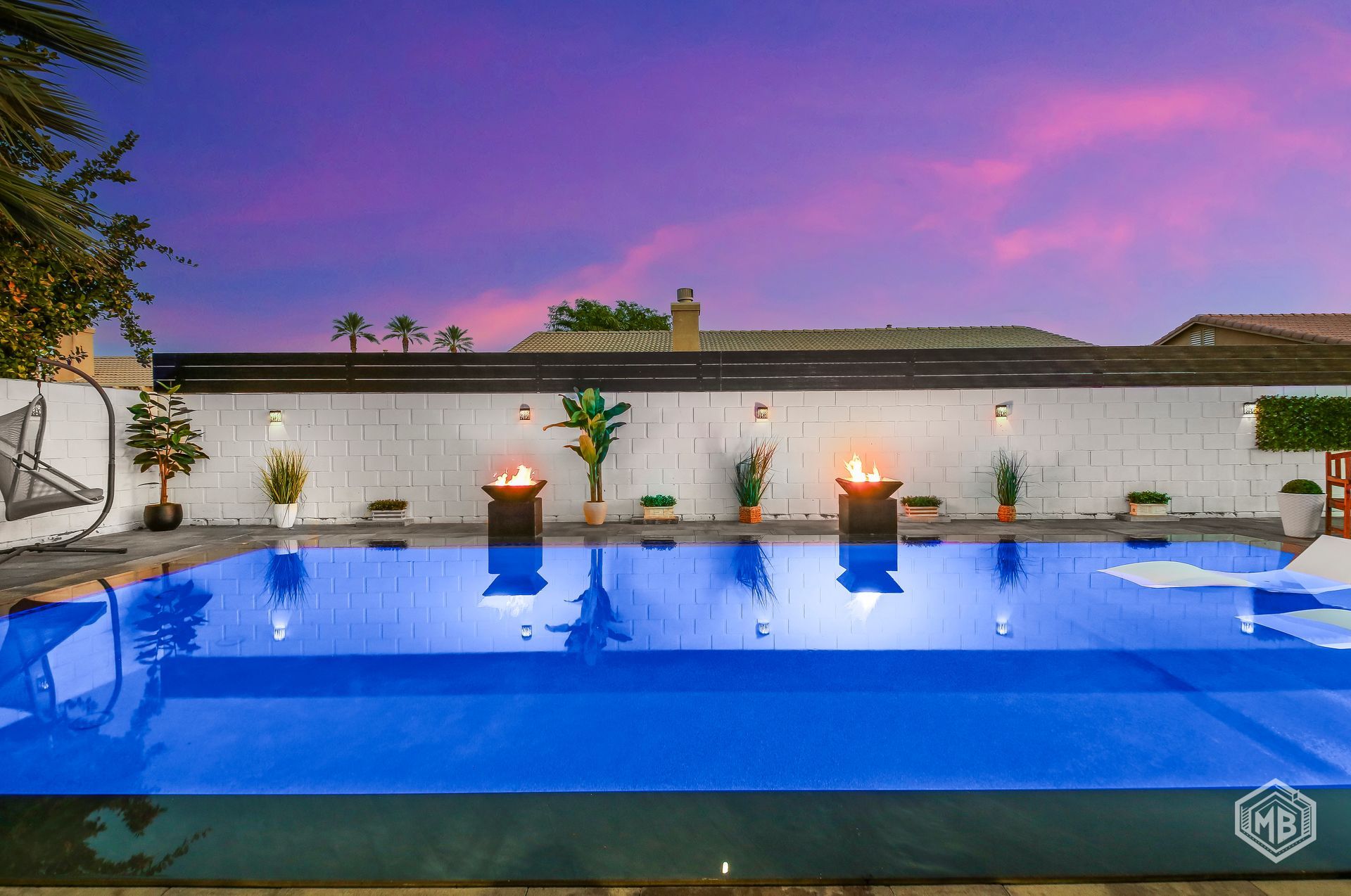 A vibrant swimming pool at twilight, flanked by glowing fire pits and potted plants against a white wall.