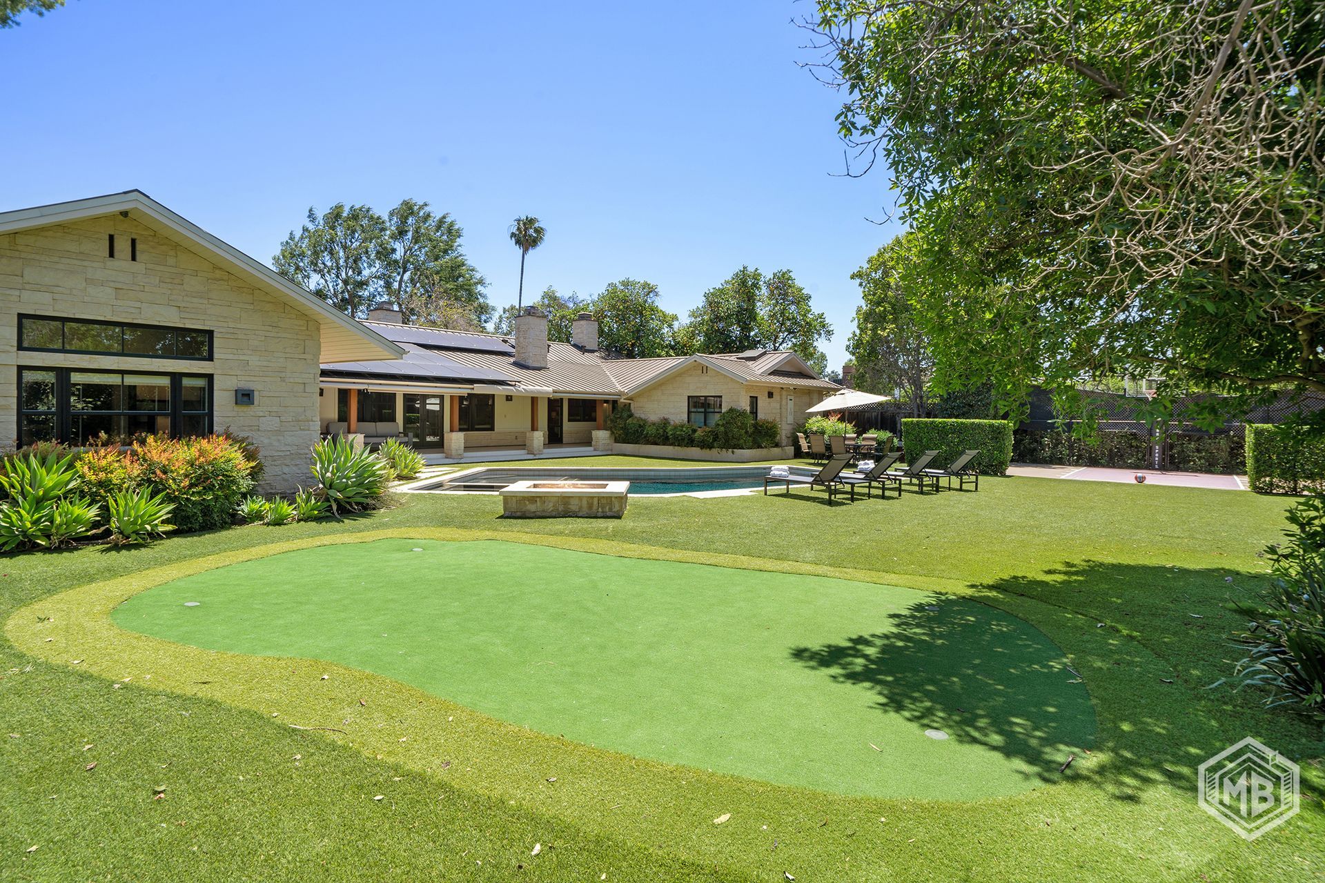 A backyard with a putting green in the foreground, a swimming pool, and a large, light-colored house under a clear sky.