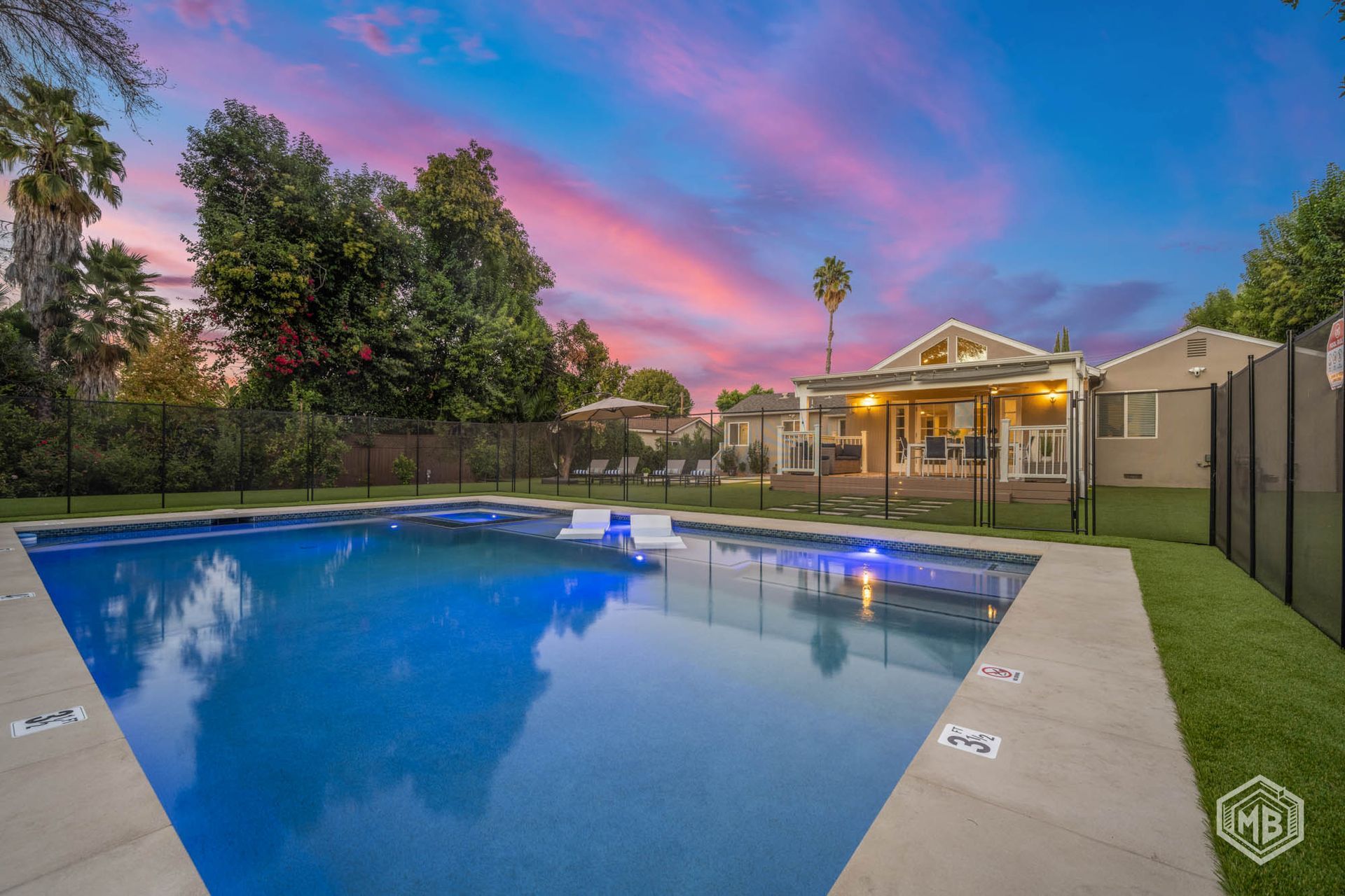 A backyard swimming pool with a patio area and lawn under a vibrant sunset sky.