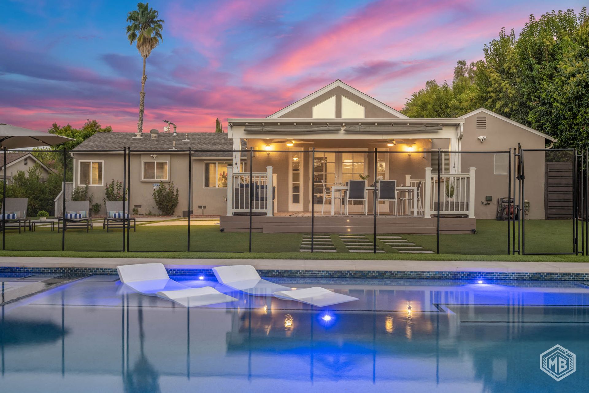 A swimming pool with lounge chairs in the foreground, facing a house with a deck at sunset with a pink and blue sky.