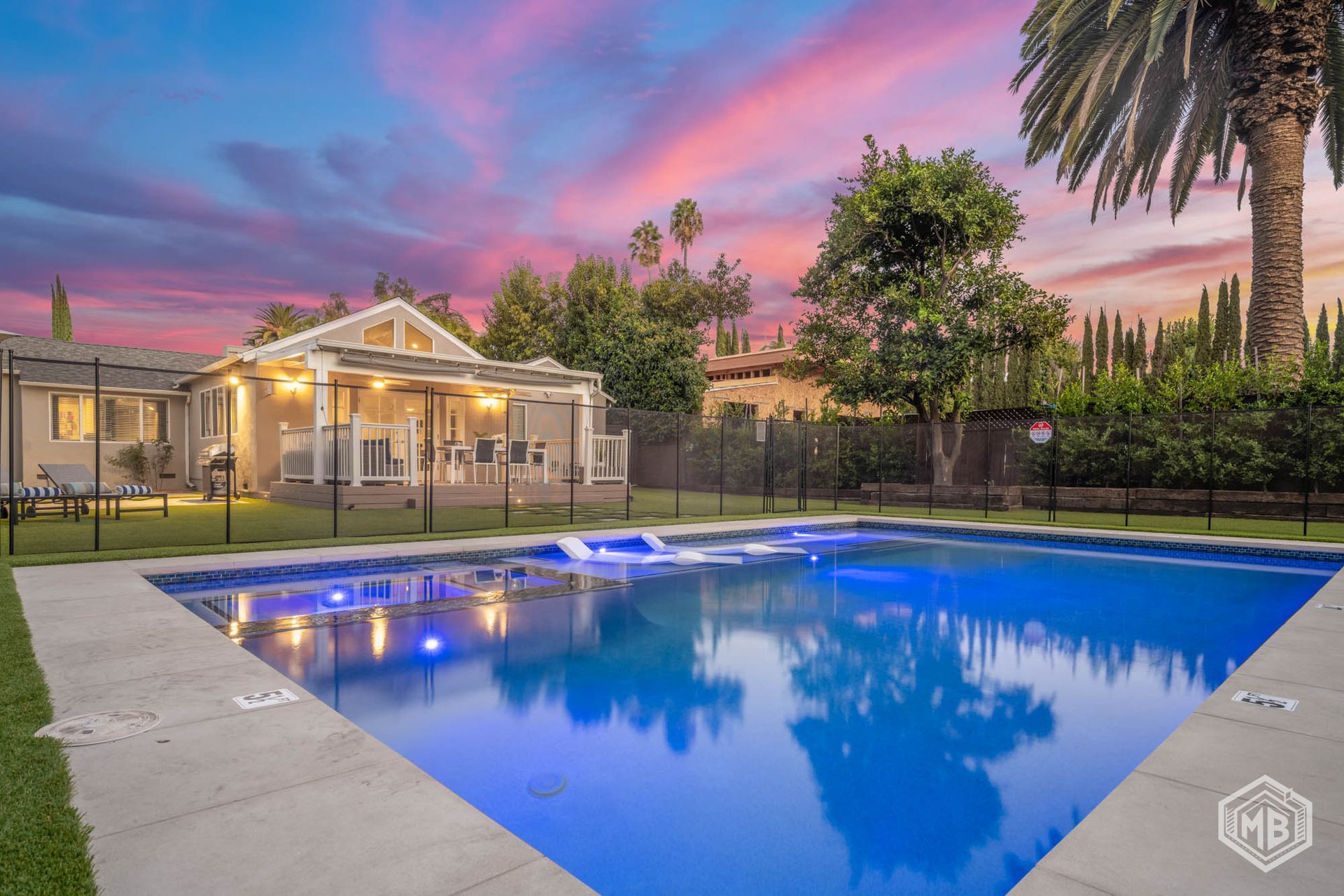 A swimming pool in a backyard with a house, patio, trees, and a vibrant sunset sky in the background.