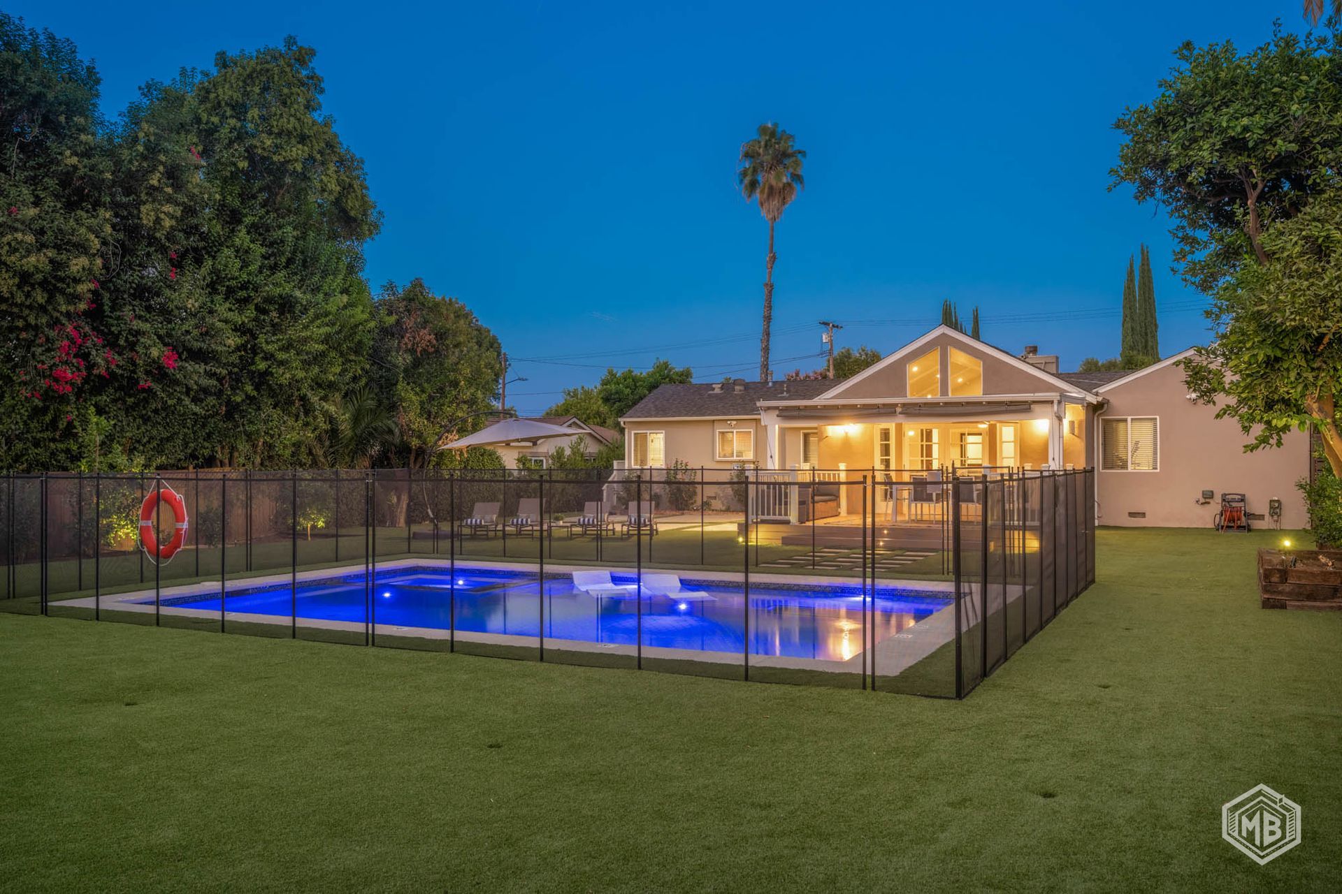 A backyard at dusk featuring a swimming pool enclosed by a black safety fence, with a lit house in the background.