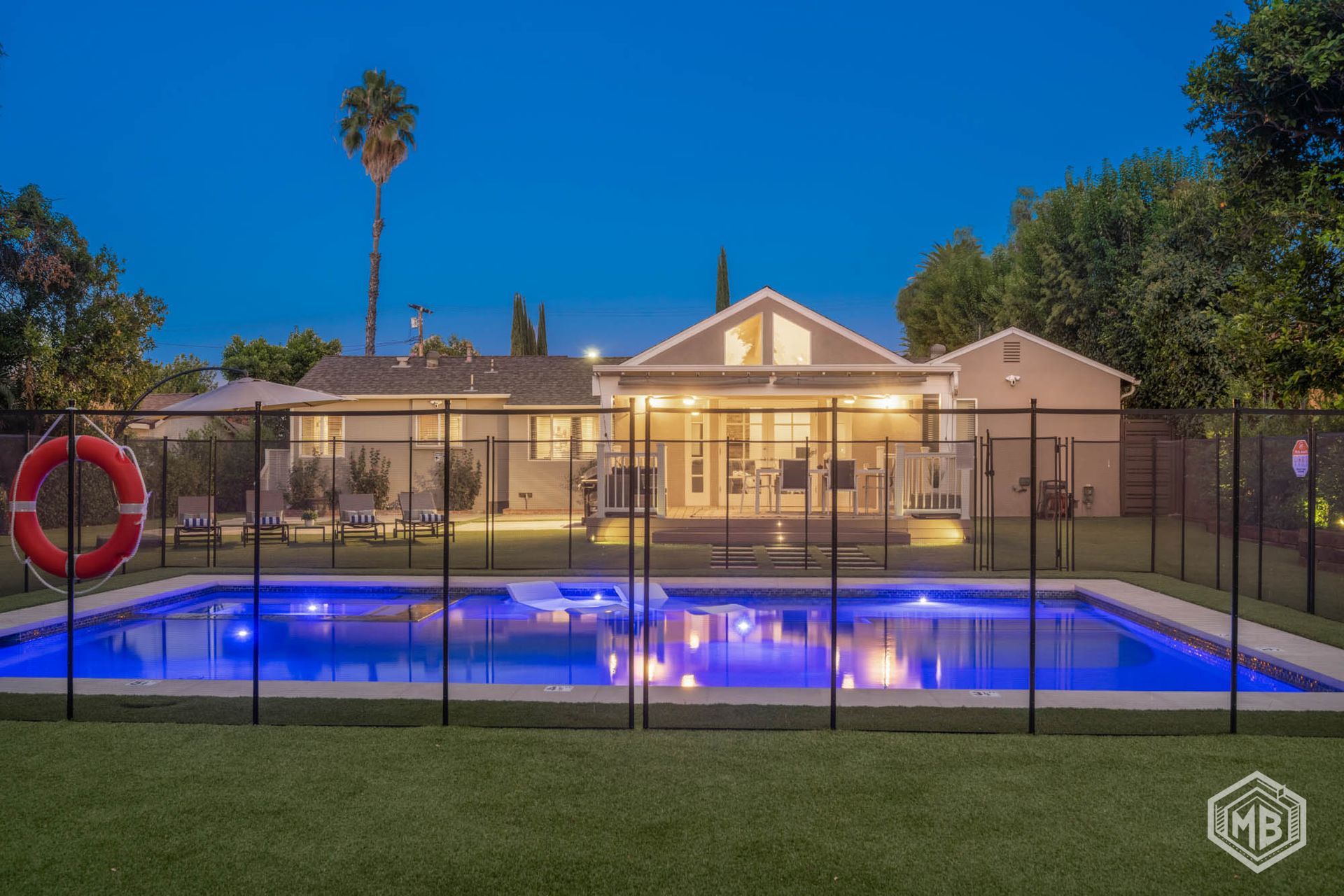 An illuminated backyard swimming pool at dusk, with a metal safety fence in the foreground and a house in the background.