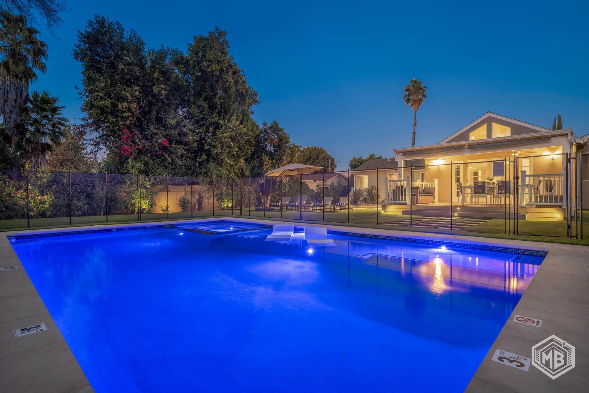 A backyard swimming pool glowing with blue light at dusk, with a lit house in the background.