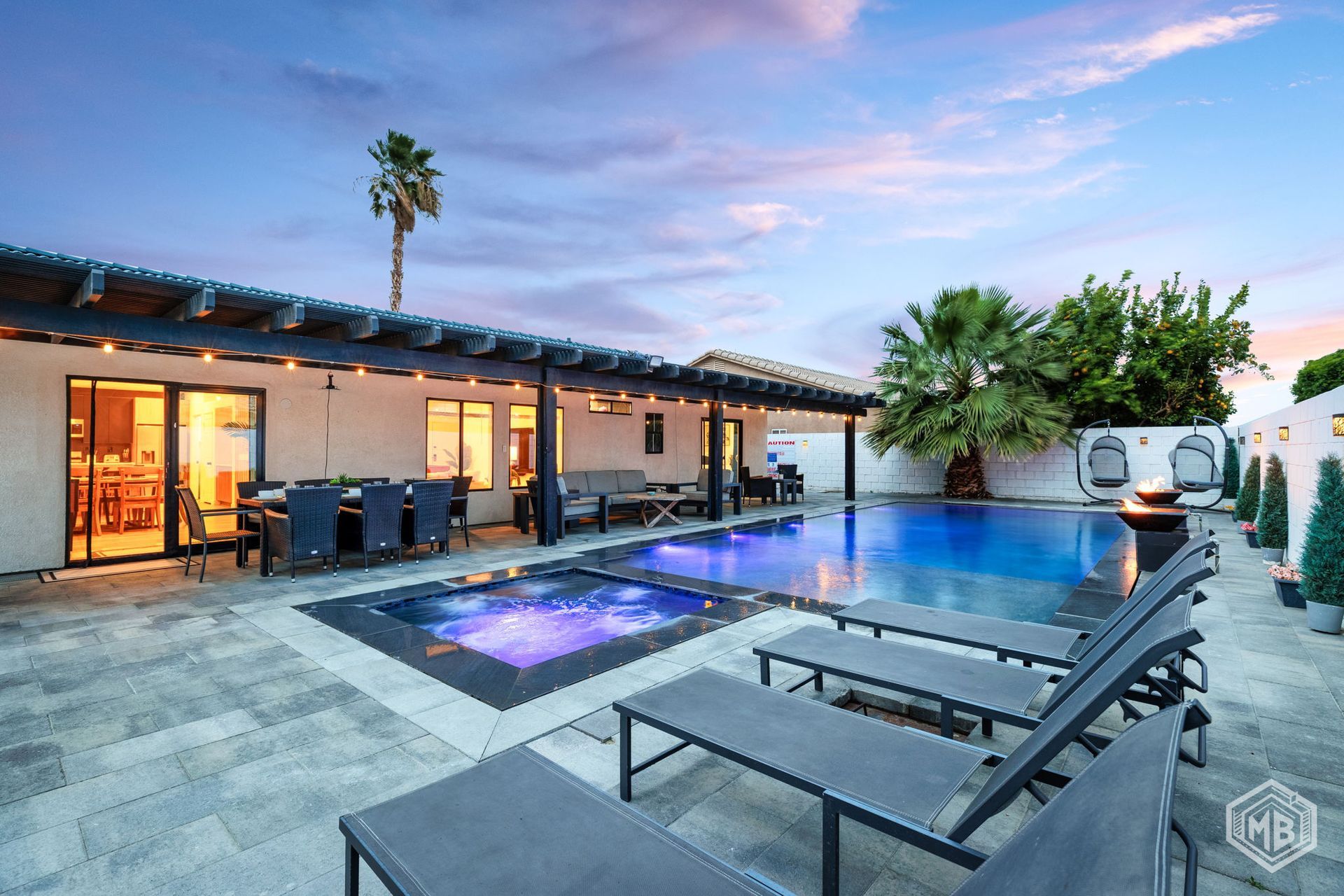 Outdoor patio with a pool and spa at dusk, featuring lounge chairs under a pergola and a lit-up backyard.