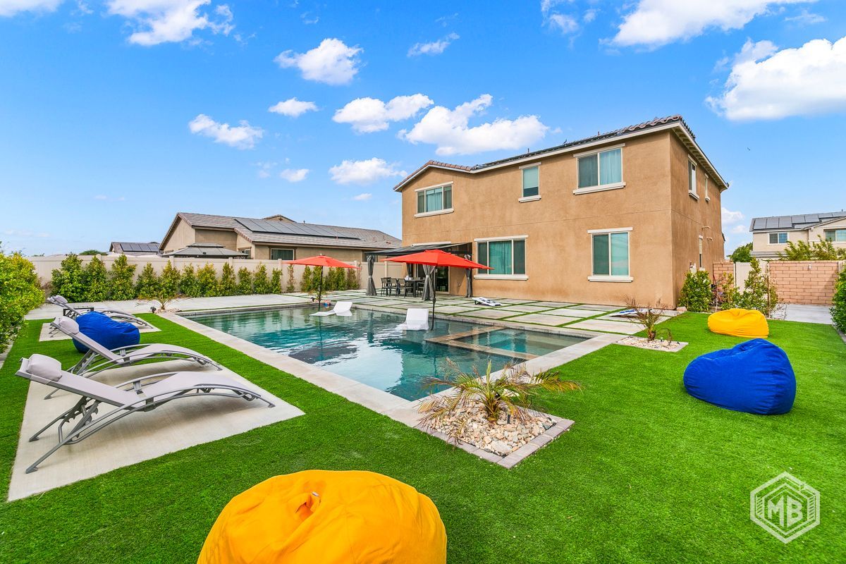 A backyard swimming pool with lounge chairs, two red umbrellas, and yellow and blue beanbag chairs on artificial grass.