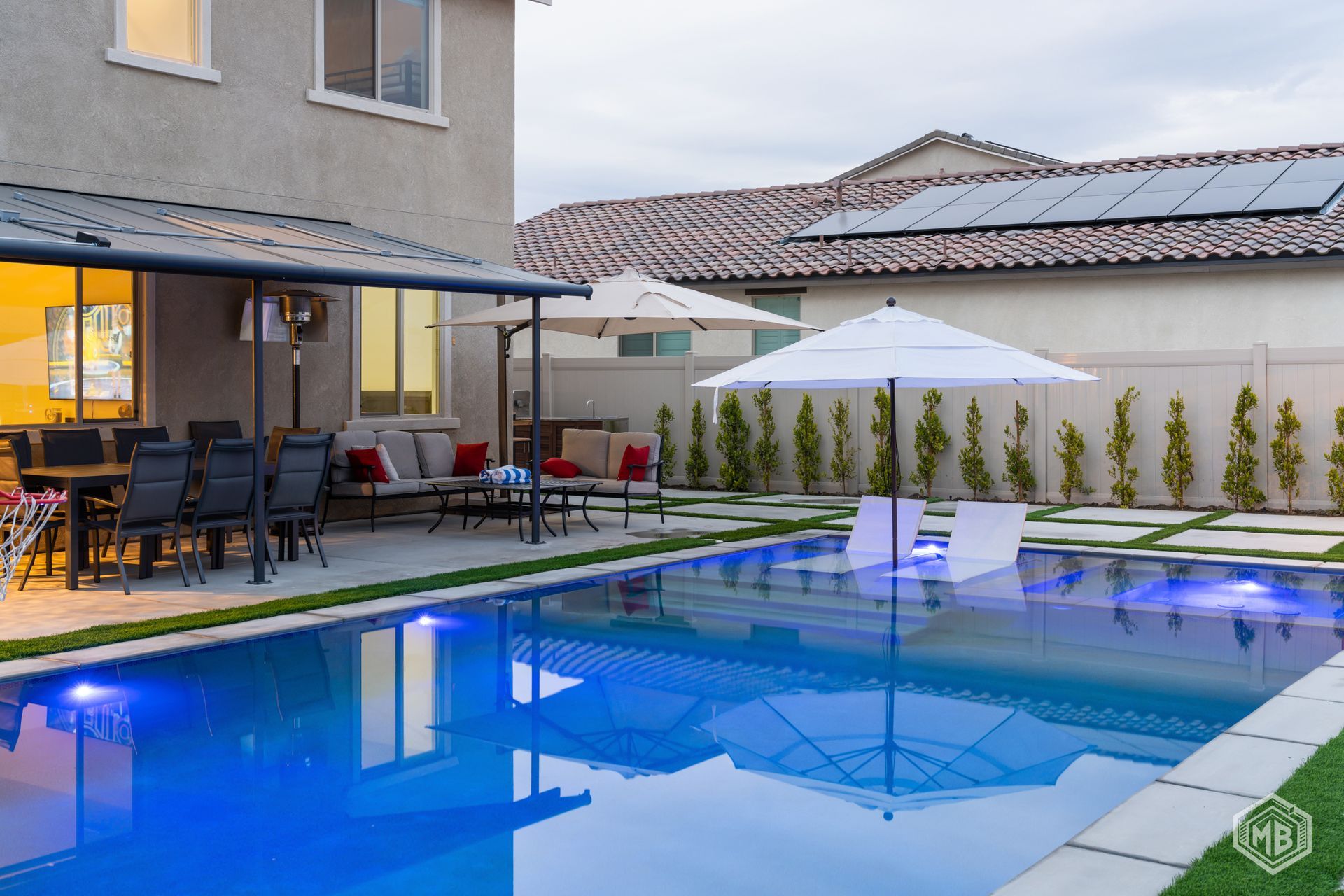 A backyard patio and pool area with a covered seating space, lounge chairs in the water, and umbrellas on a sunny day.