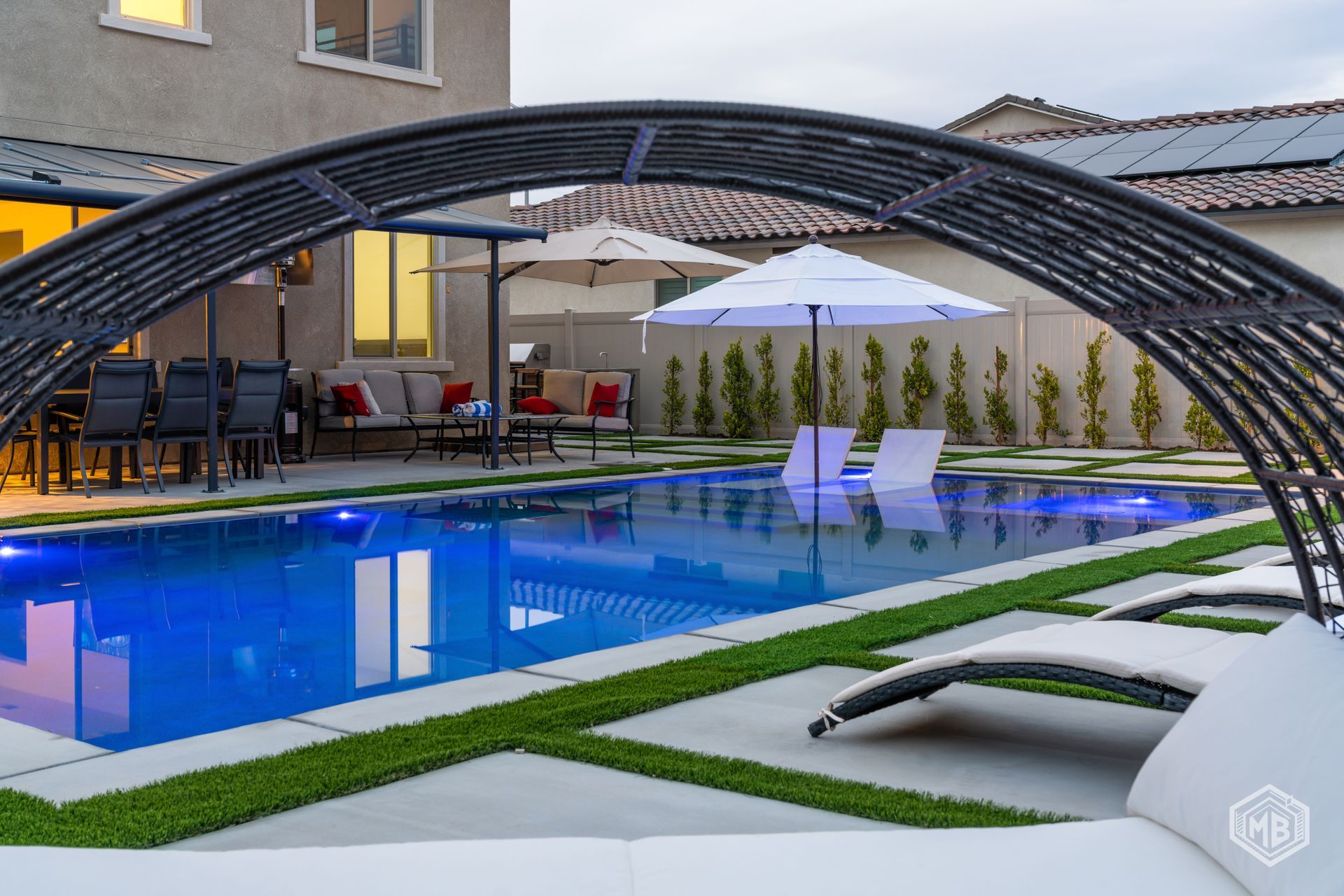 A modern backyard with a blue swimming pool, patio furniture, loungers, and an arched shade structure.