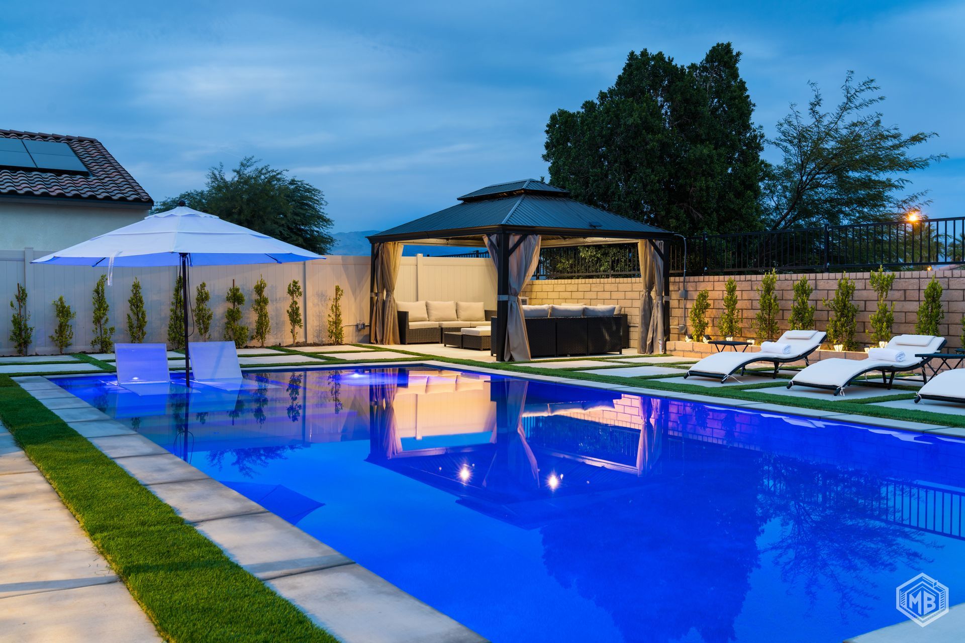 A blue pool at twilight with a white umbrella, poolside lounge chairs, and a covered patio structure in a backyard.