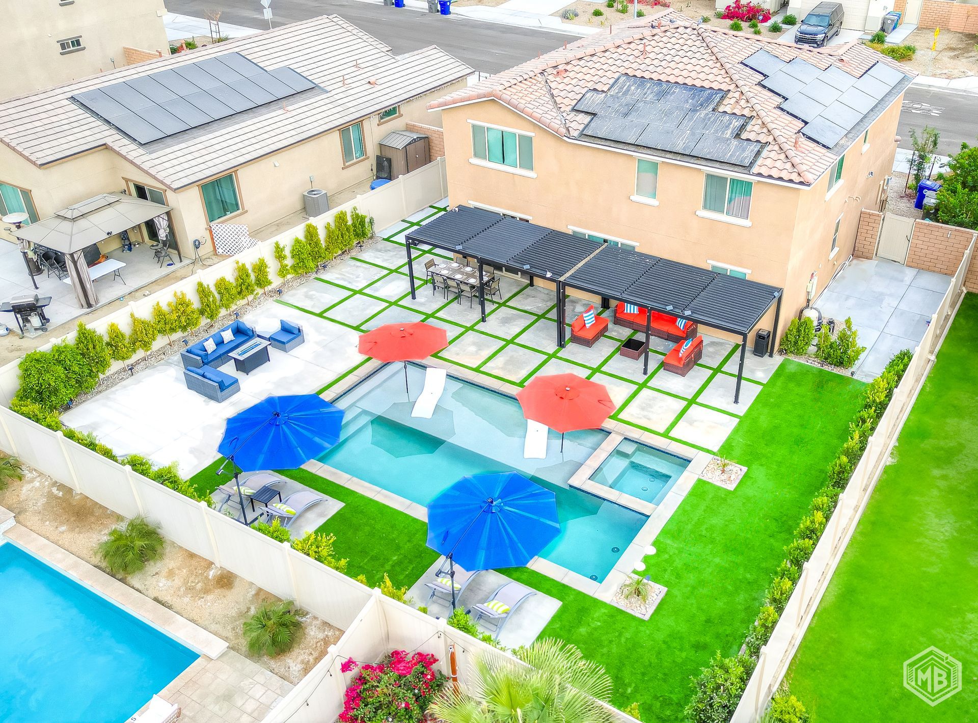 A backyard with a swimming pool, spa, covered patio with outdoor furniture, and red and blue umbrellas on artificial turf.