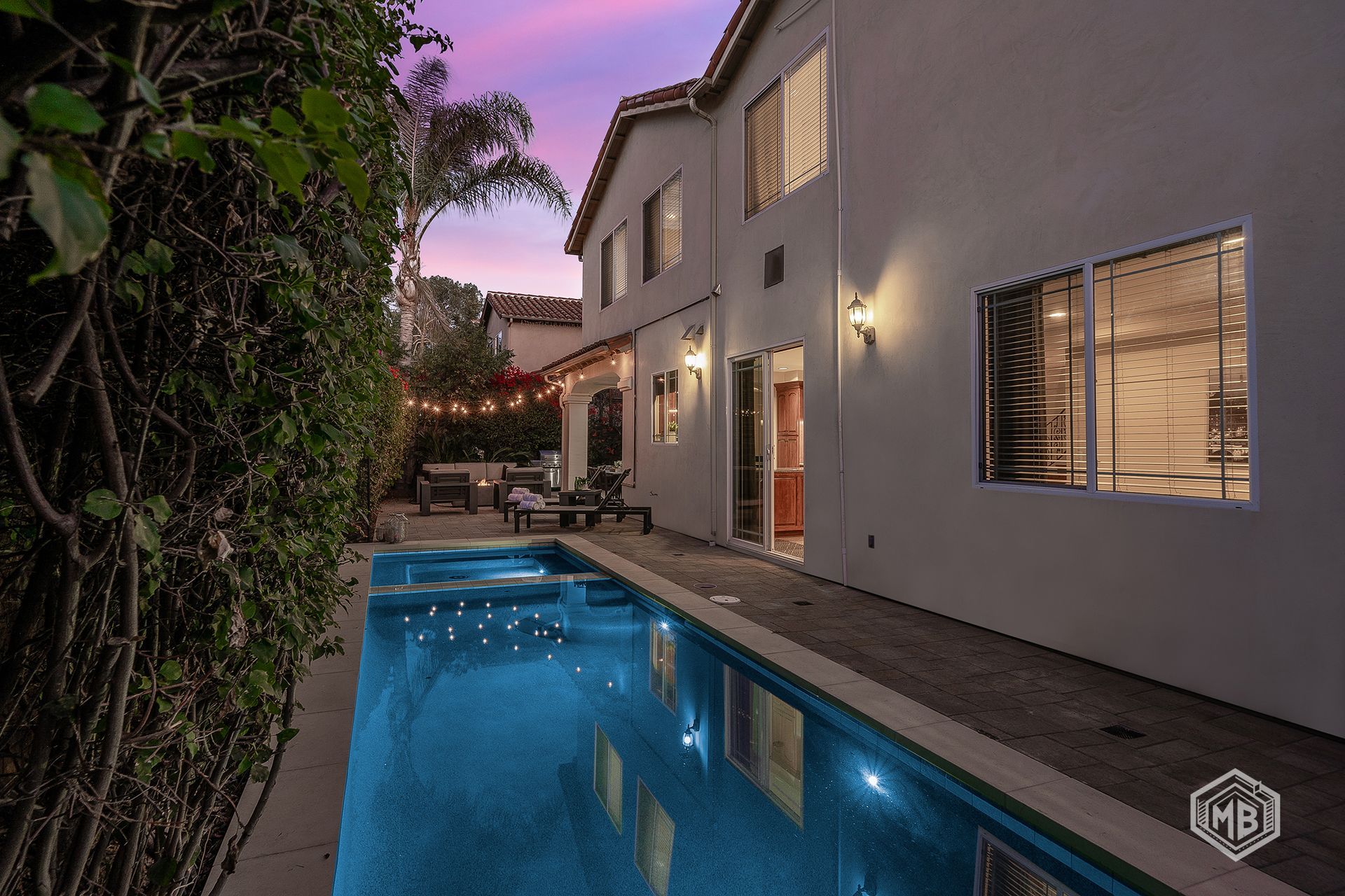 A backyard with a long rectangular pool and spa, lit by evening string lights against a purple and orange sunset sky.