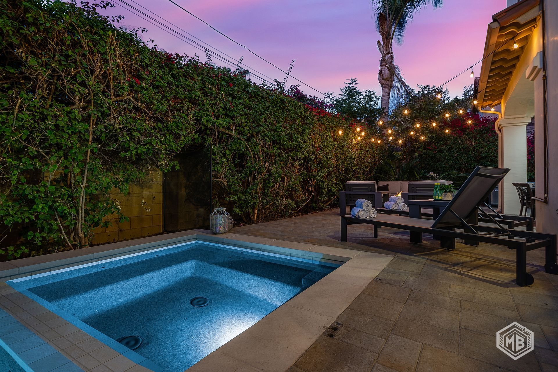 A small illuminated pool on a stone patio at dusk, featuring lounge chairs under string lights against a lush hedge.