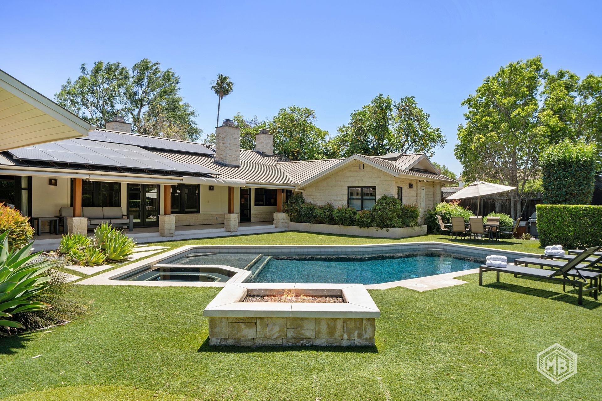 A backyard with a pool, a stone fire pit, and a house with a patio, surrounded by green grass and trees under a clear sky.