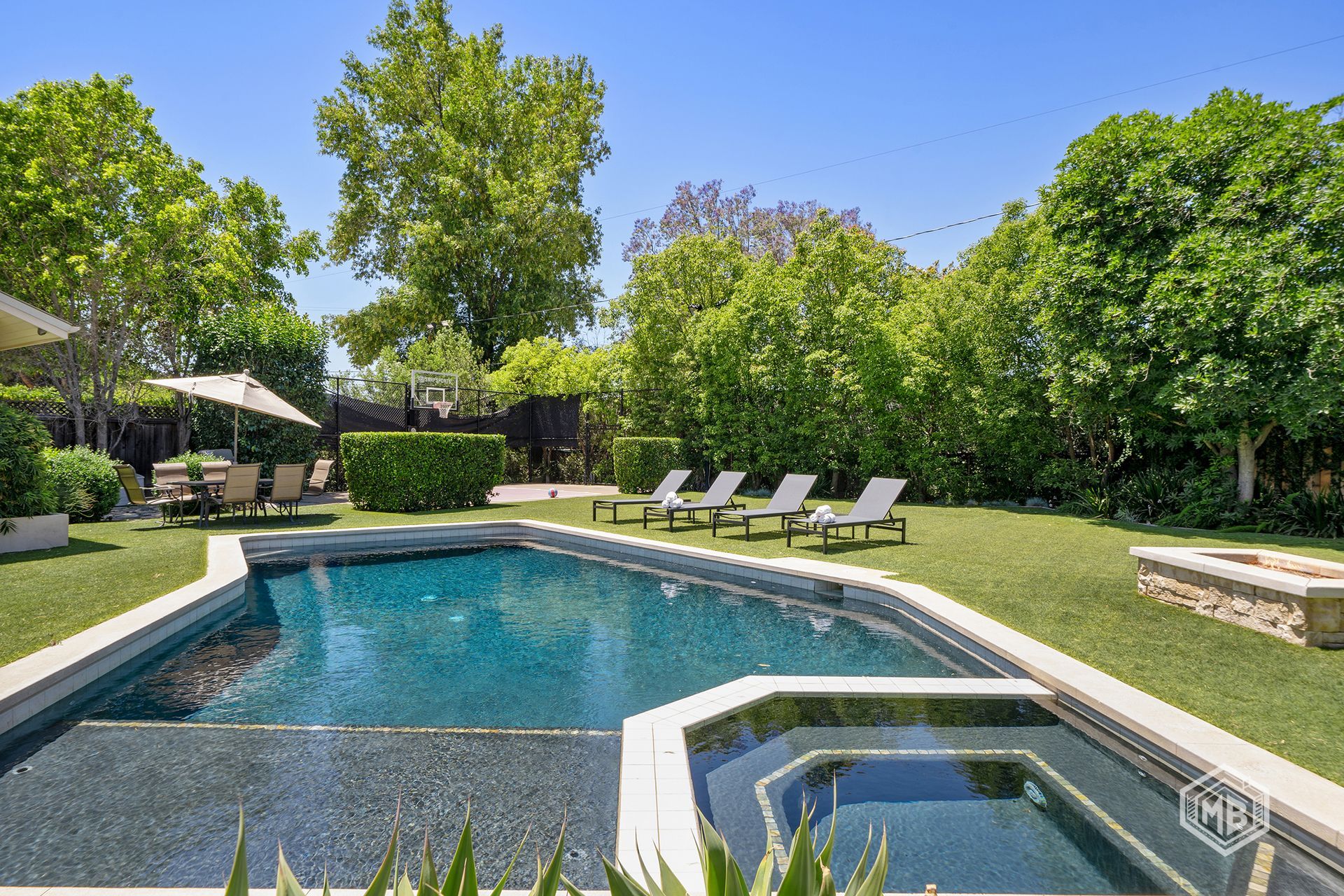 A backyard pool and hot tub surrounded by a lawn, patio furniture, and lush green trees under a bright blue sky.