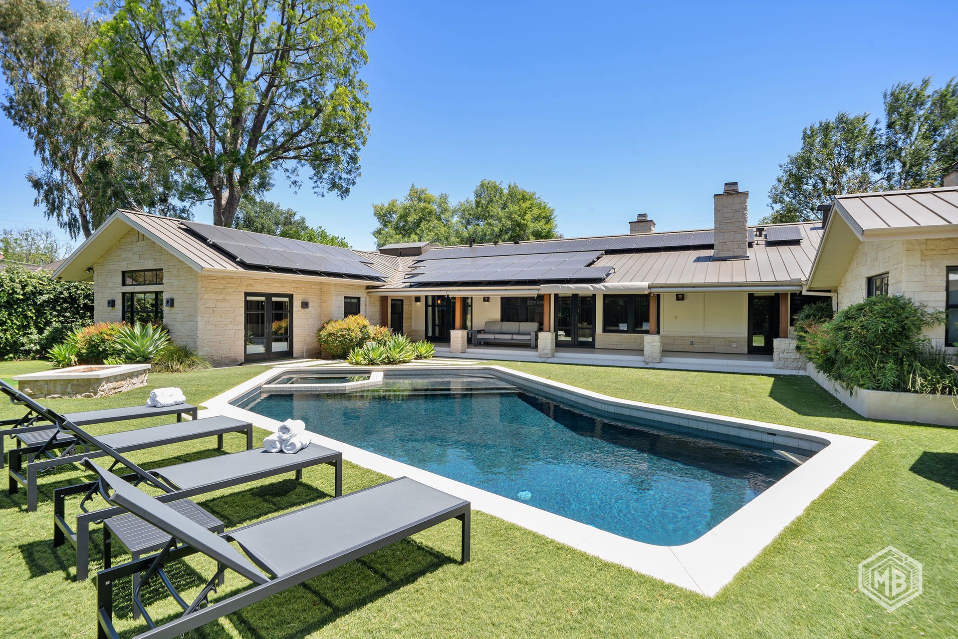 A backyard with a swimming pool, lounge chairs on green grass, and a single-story stone house with a metal roof.