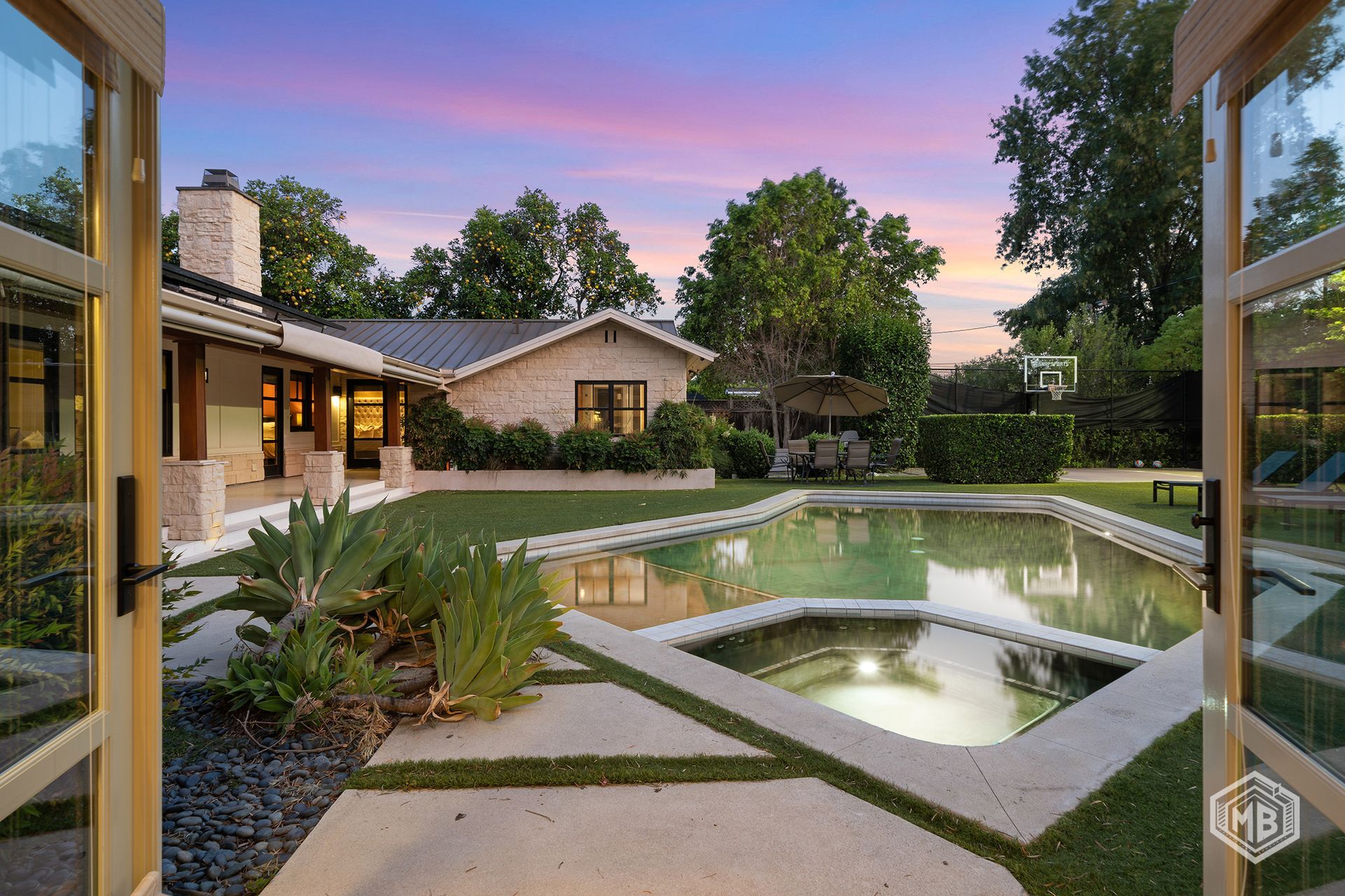 A backyard pool and hot tub at dusk, viewed through open French doors from inside a house with light stone architecture.