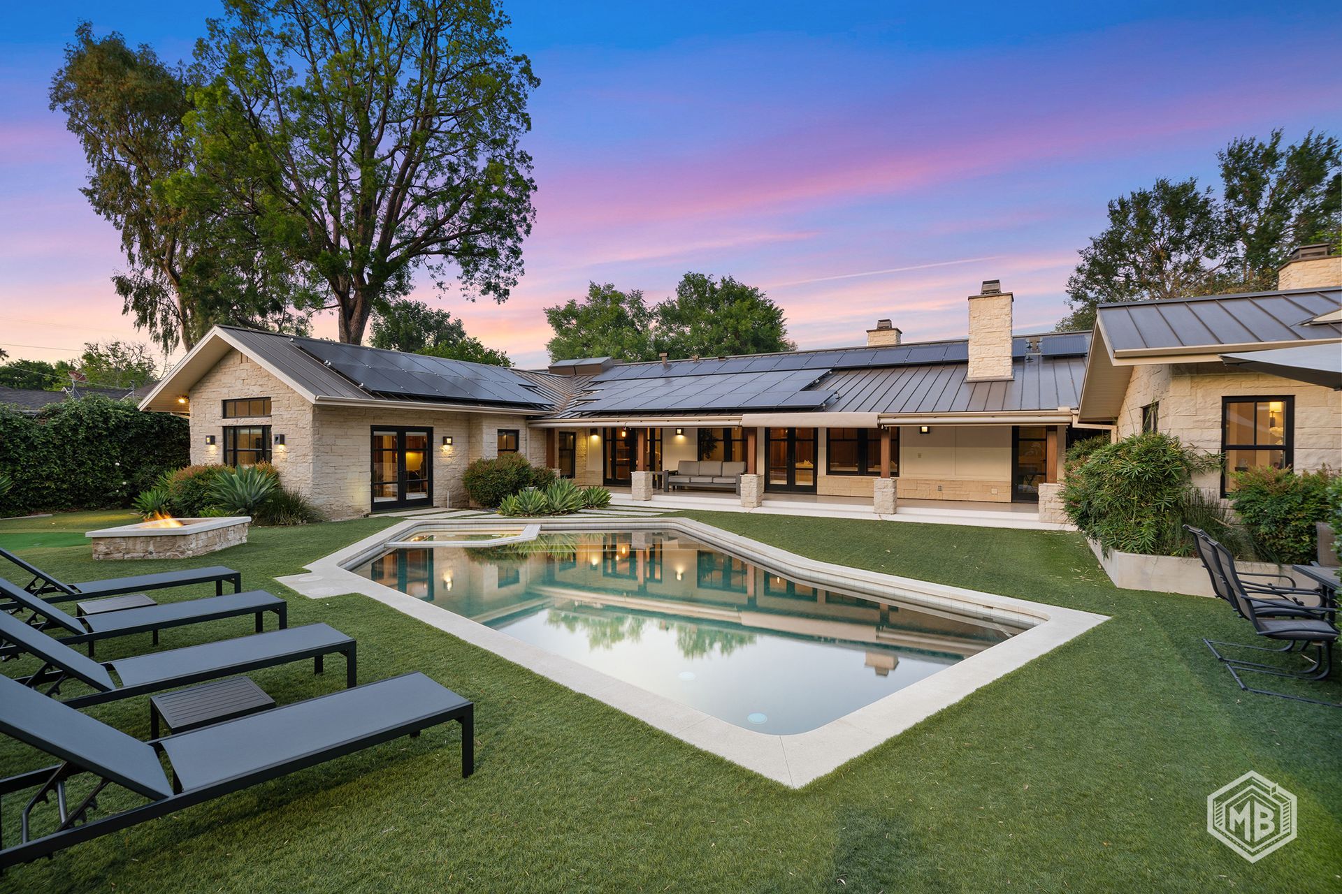 A modern, one-story stone house with a metal roof overlooks a backyard pool at twilight under a pink and blue sky.