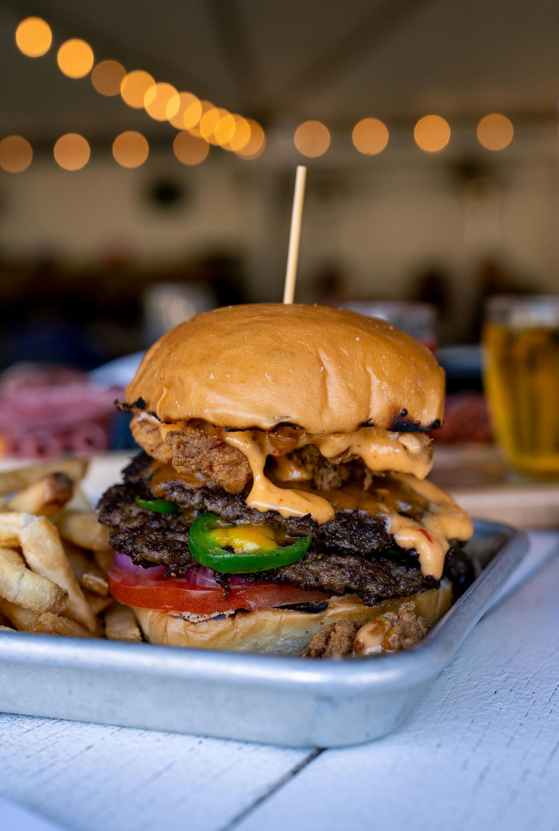 A double patty burger topped with jalapeños and sauce, served with fries on a metal tray with blurry lights background.