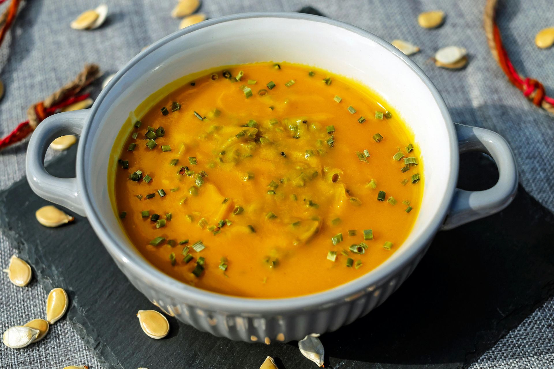 A grey bowl filled with orange pumpkin soup, garnished with herbs, on a dark slate mat with scattered pumpkin seeds.