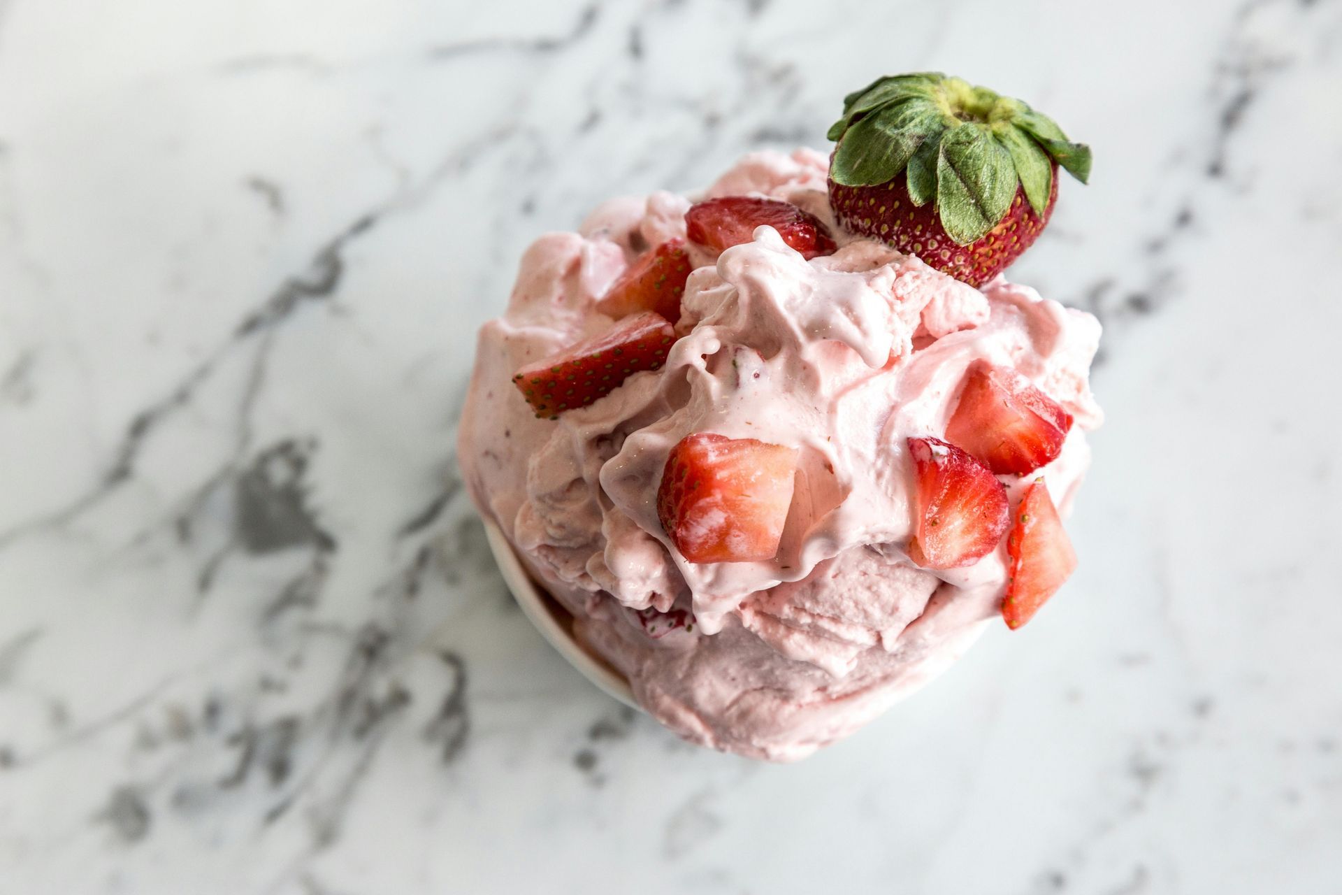 A white bowl filled with strawberry mousse or ice cream, topped with fresh strawberry slices, on a white marble surface.