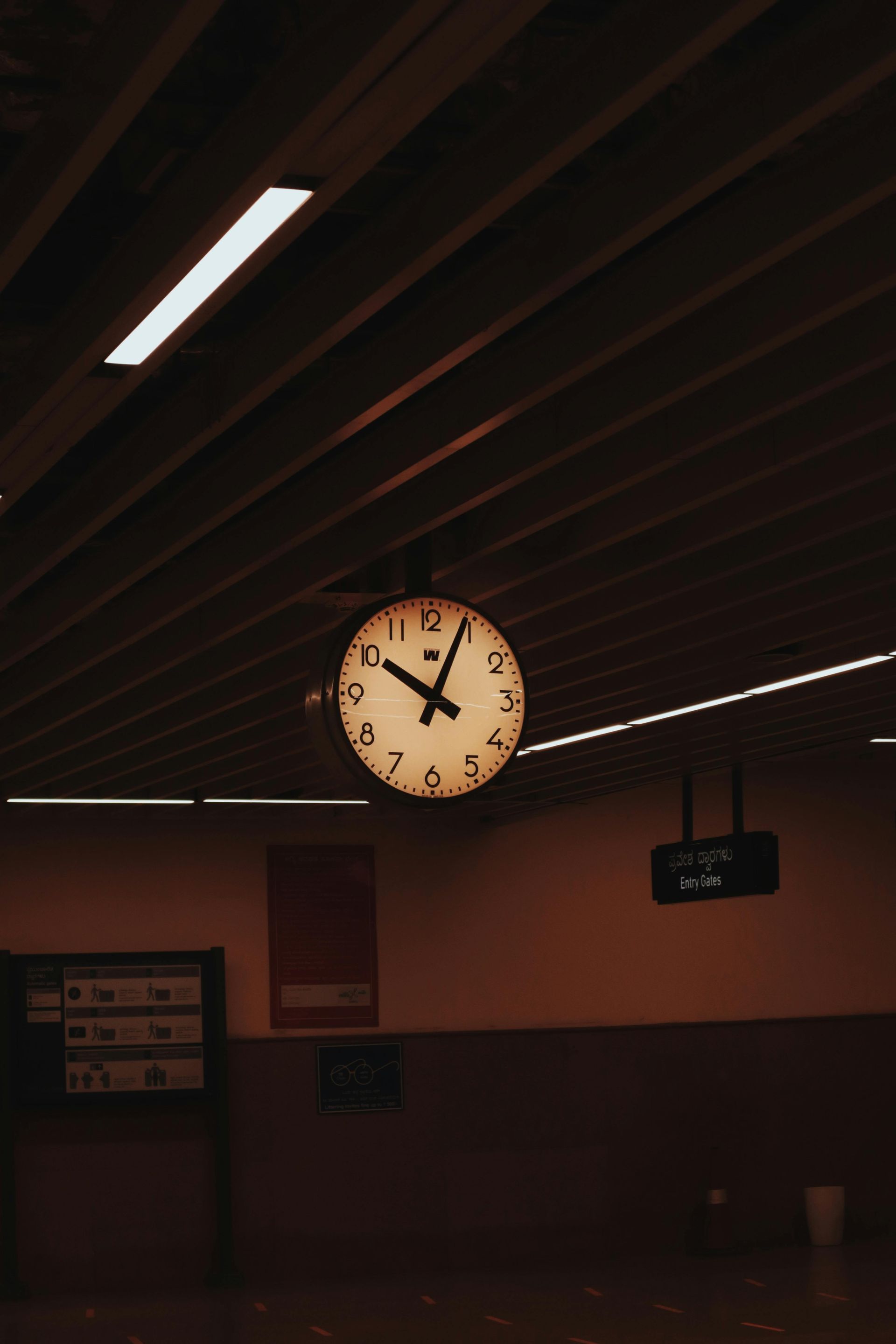 A round clock hangs from a slatted ceiling in a dimly lit, modern subway station platform.