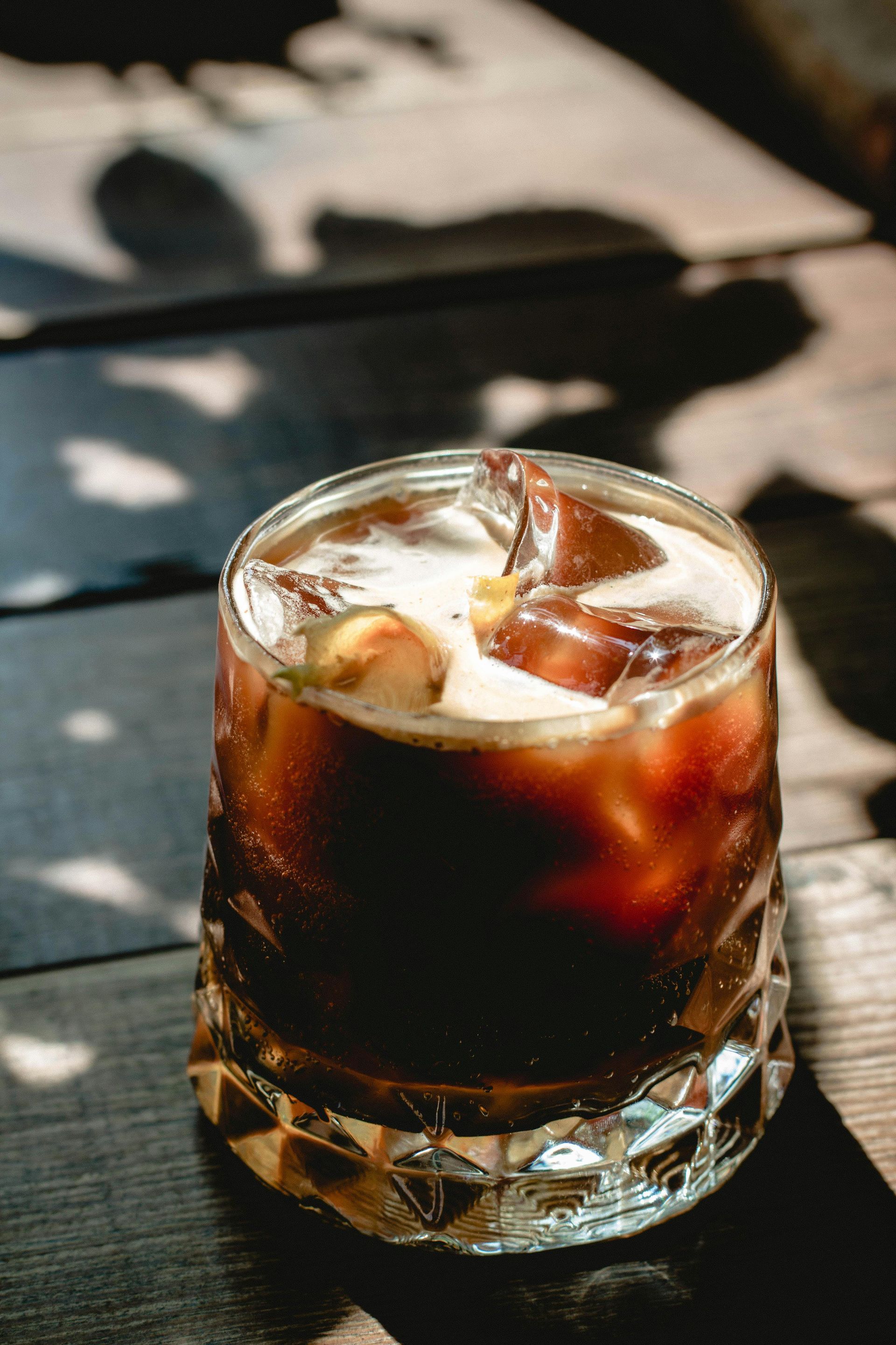A glass of iced coffee in a faceted tumbler, sitting on a dark wooden table with soft, dappled sunlight shadows.