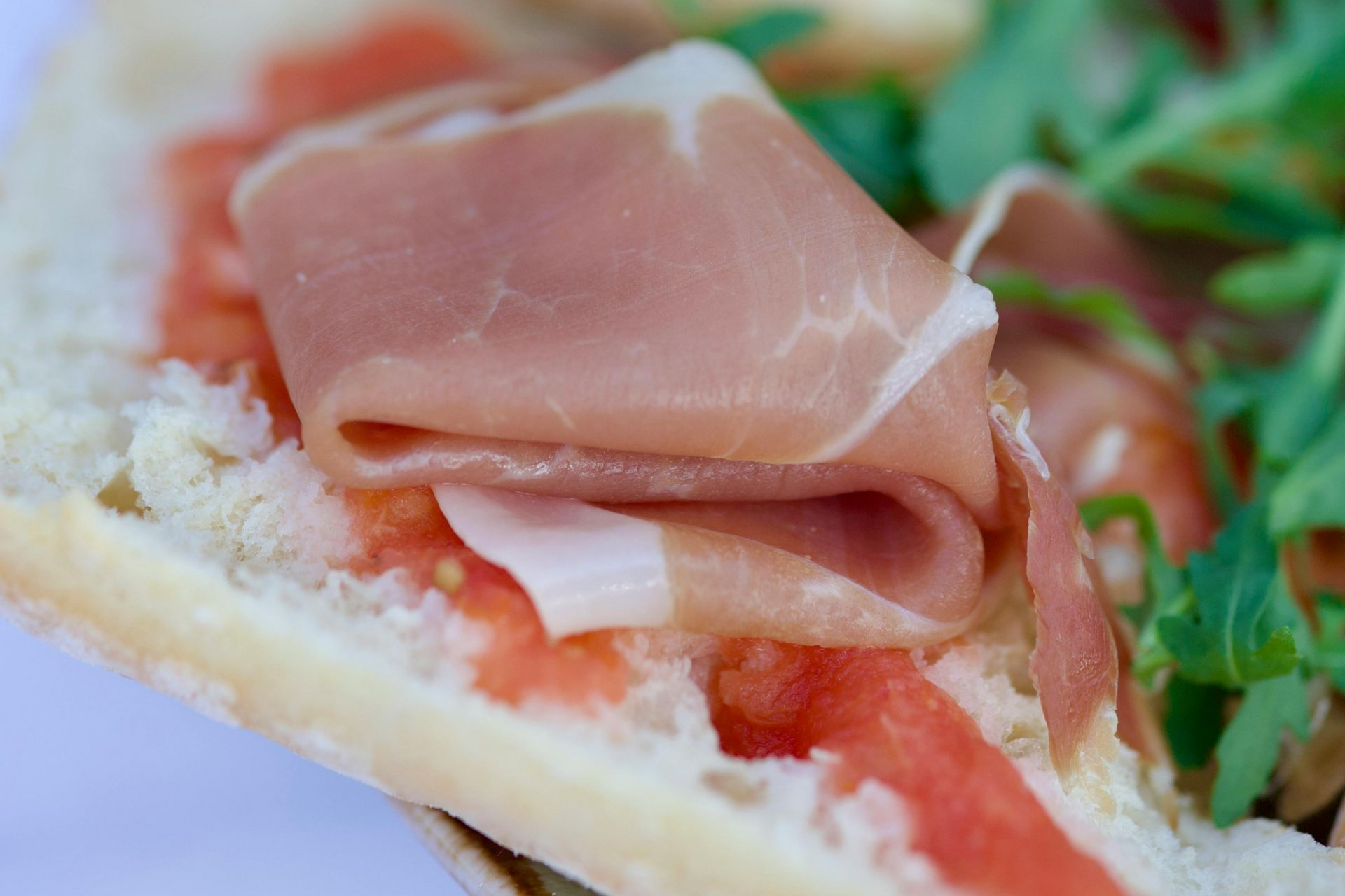 A close-up of a crusty bread slice topped with tomato sauce, folded slices of prosciutto, and fresh arugula.