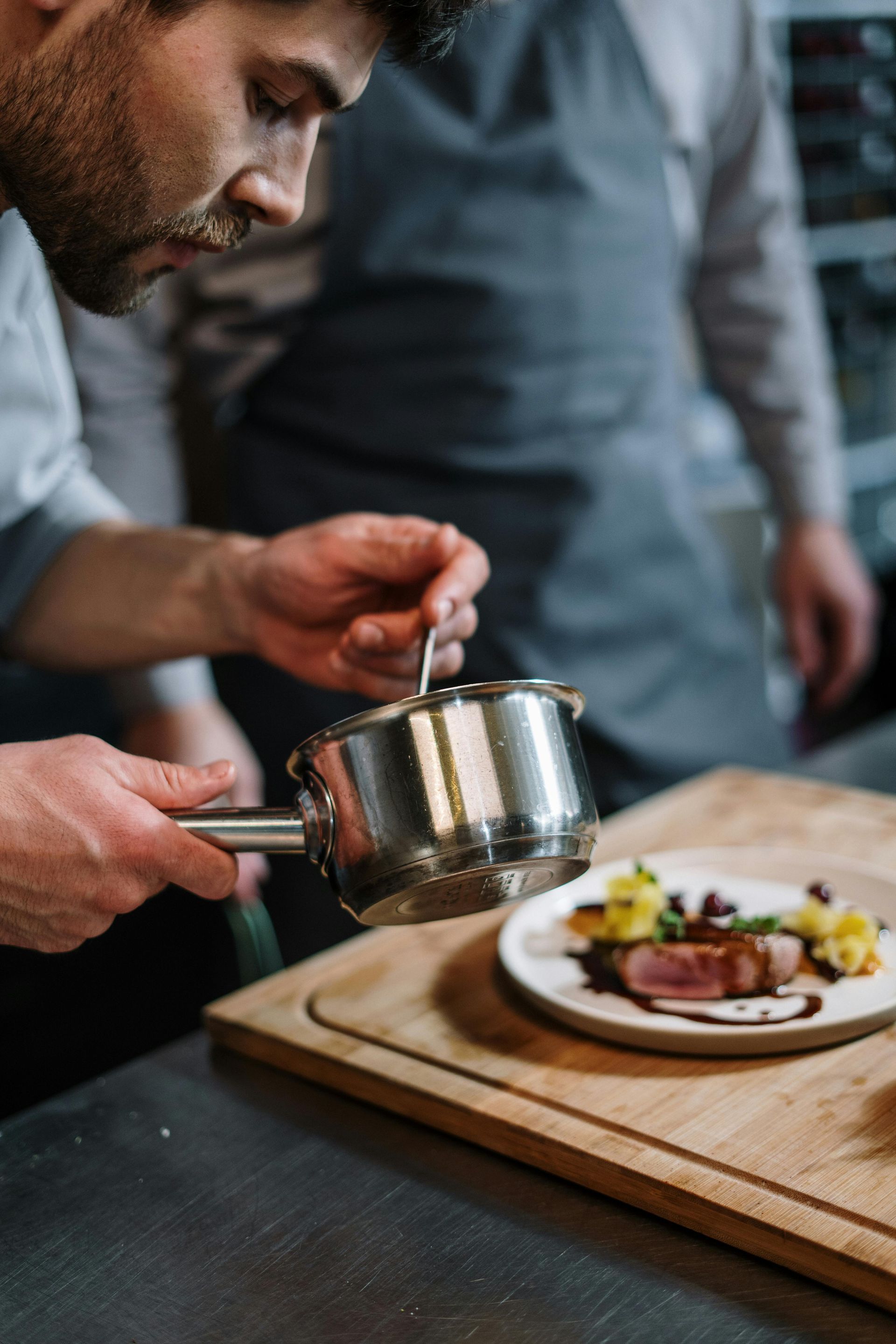 A person in a kitchen uses a small metal saucepan and spoon to drizzle sauce onto a plated meal on a wooden cutting board.
