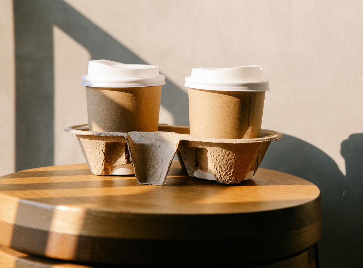 Two tan paper coffee cups in a cardboard carrier, sitting on a round wooden surface in warm sunlight.