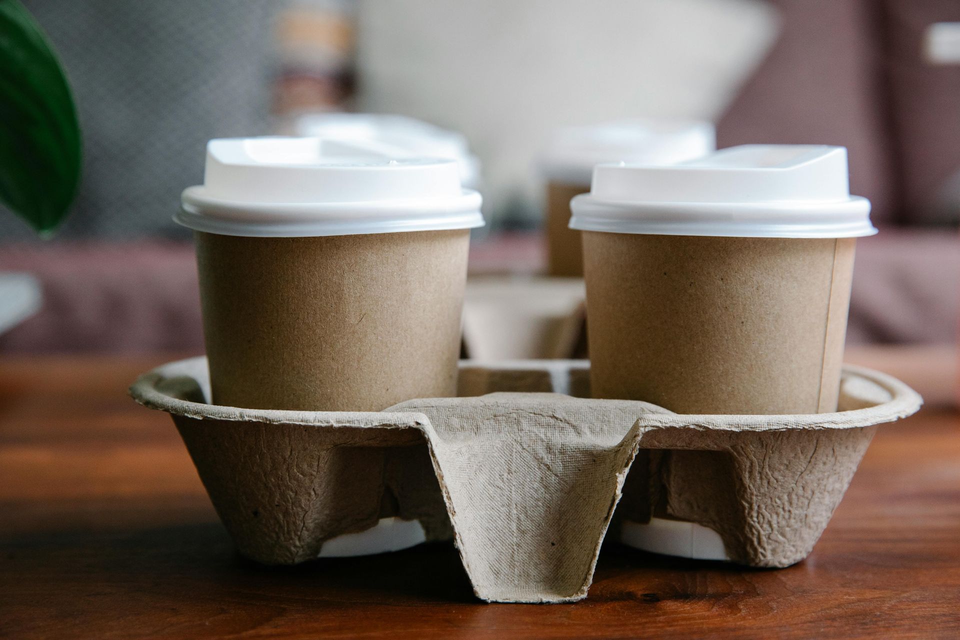 Two beige disposable coffee cups with white lids, sitting in a molded fiber carrier on a wooden table.