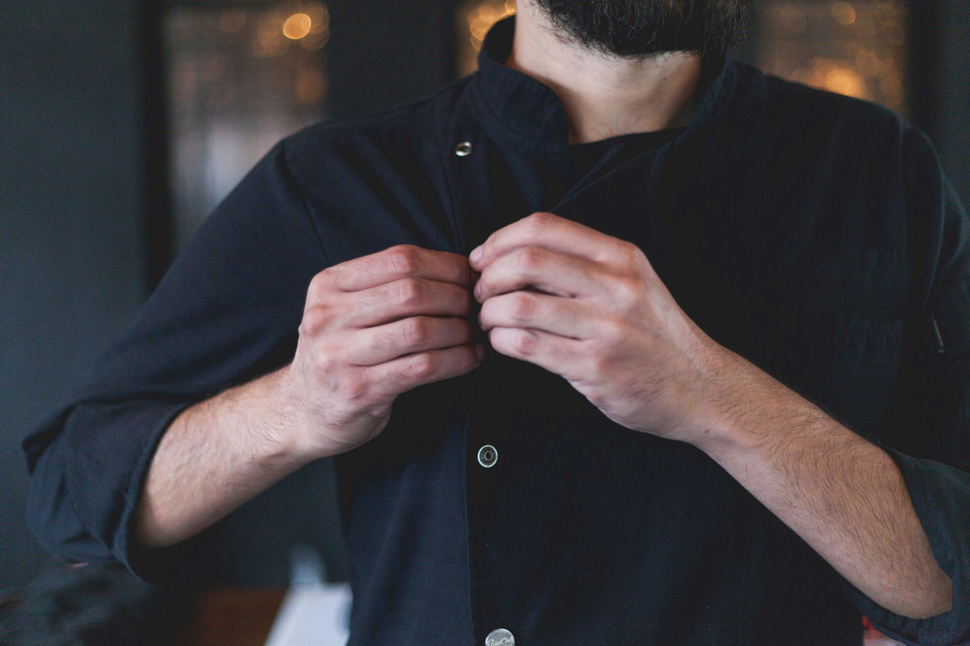 A person with a beard buttons a dark, professional chef's coat, with hands in focus near the center.