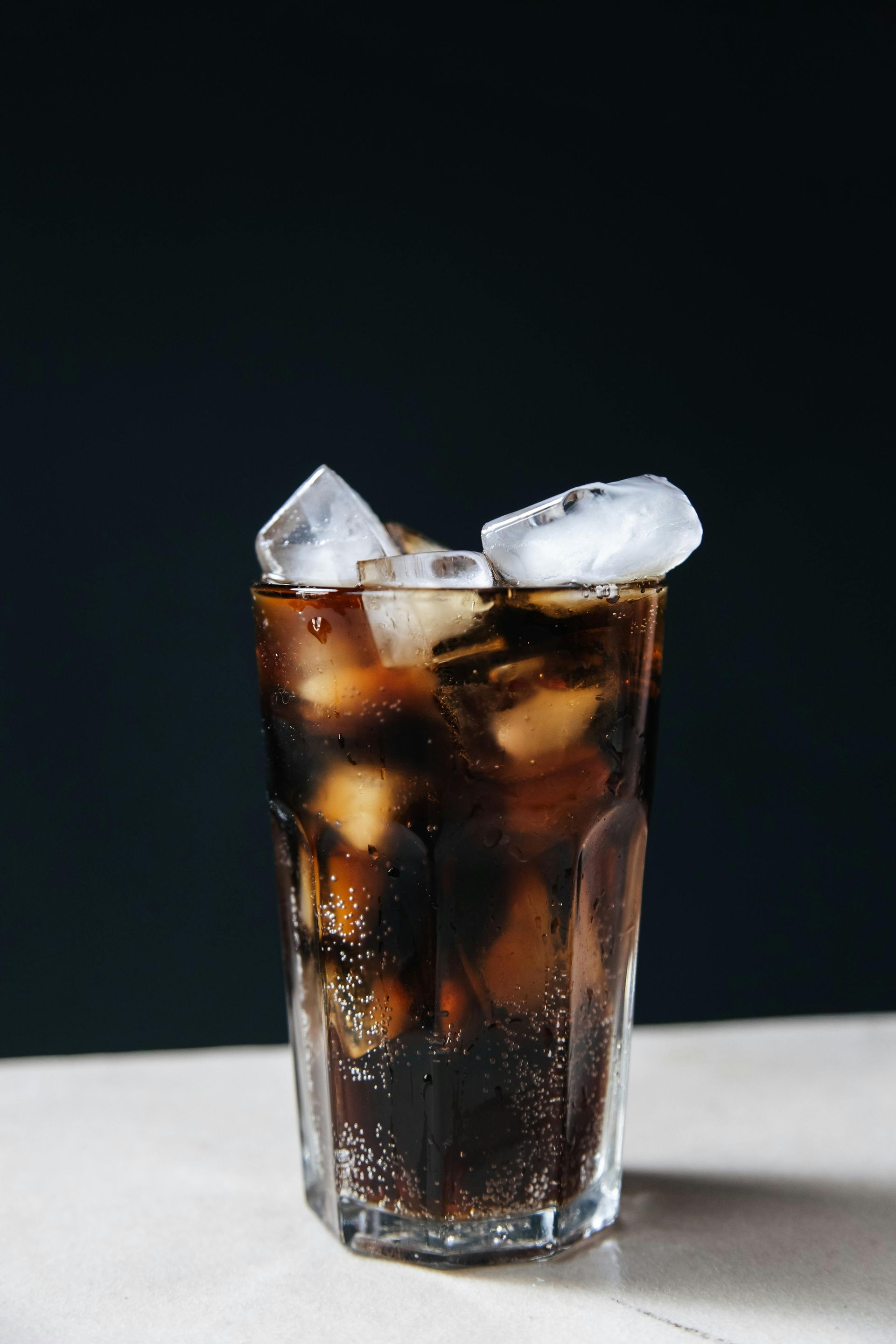 A clear glass filled with dark, carbonated soda and ice cubes, resting on a white surface against a dark background.