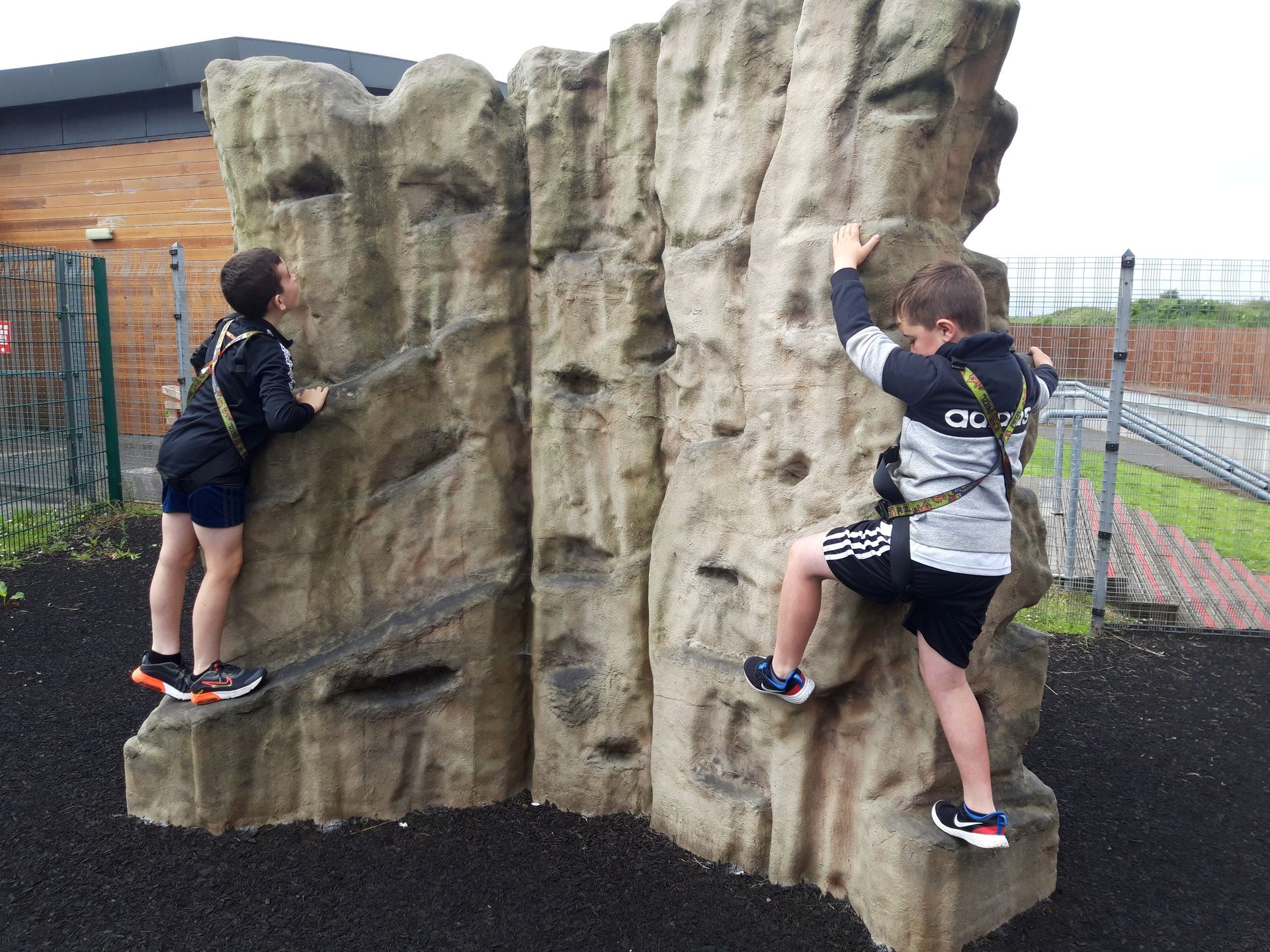 Two children climbing a rock wall outside. One has arms raised, other is holding on. Black safety harnesses.