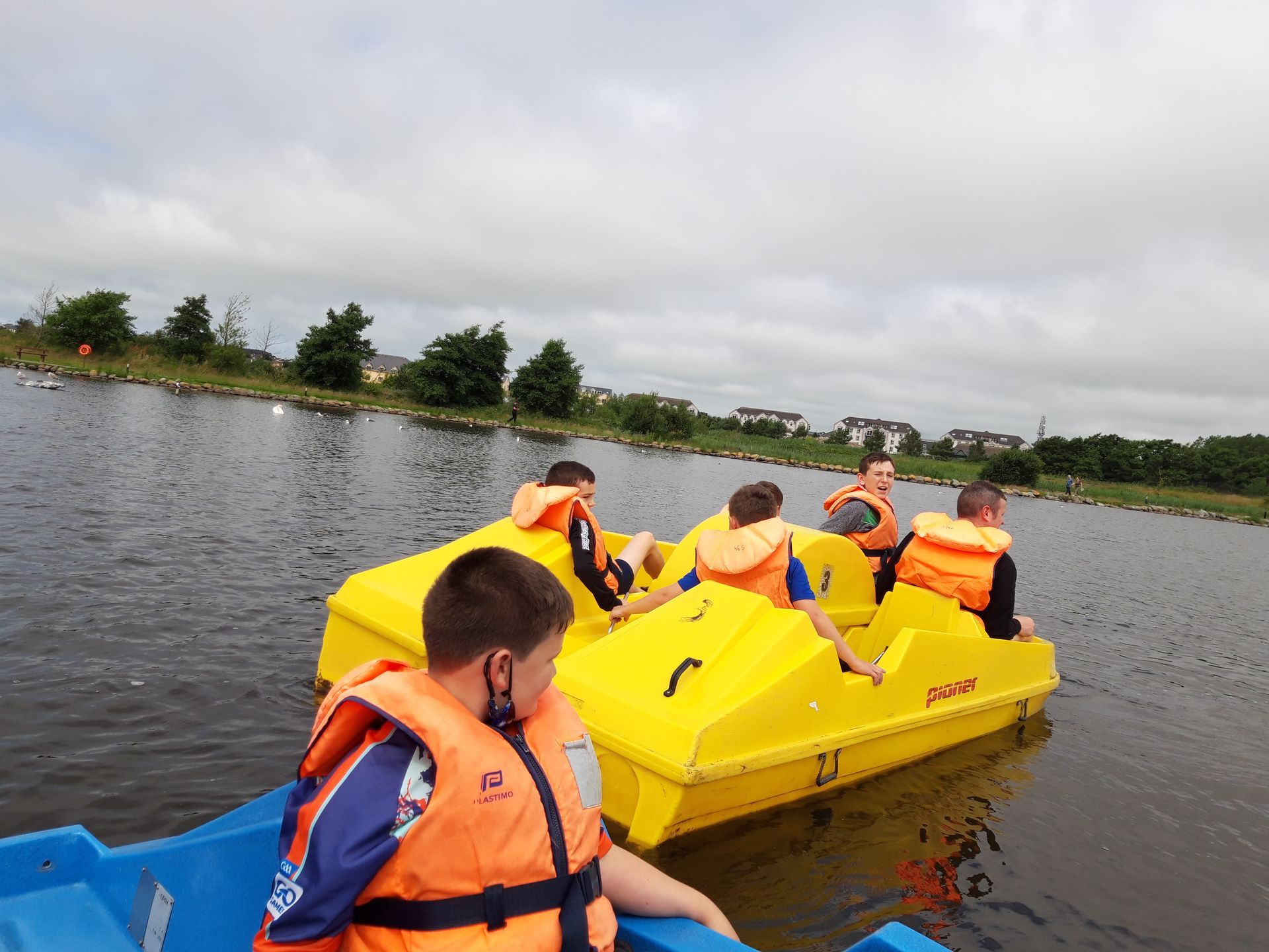 People in yellow paddle boats on a lake, wearing orange life vests. Cloudy sky.