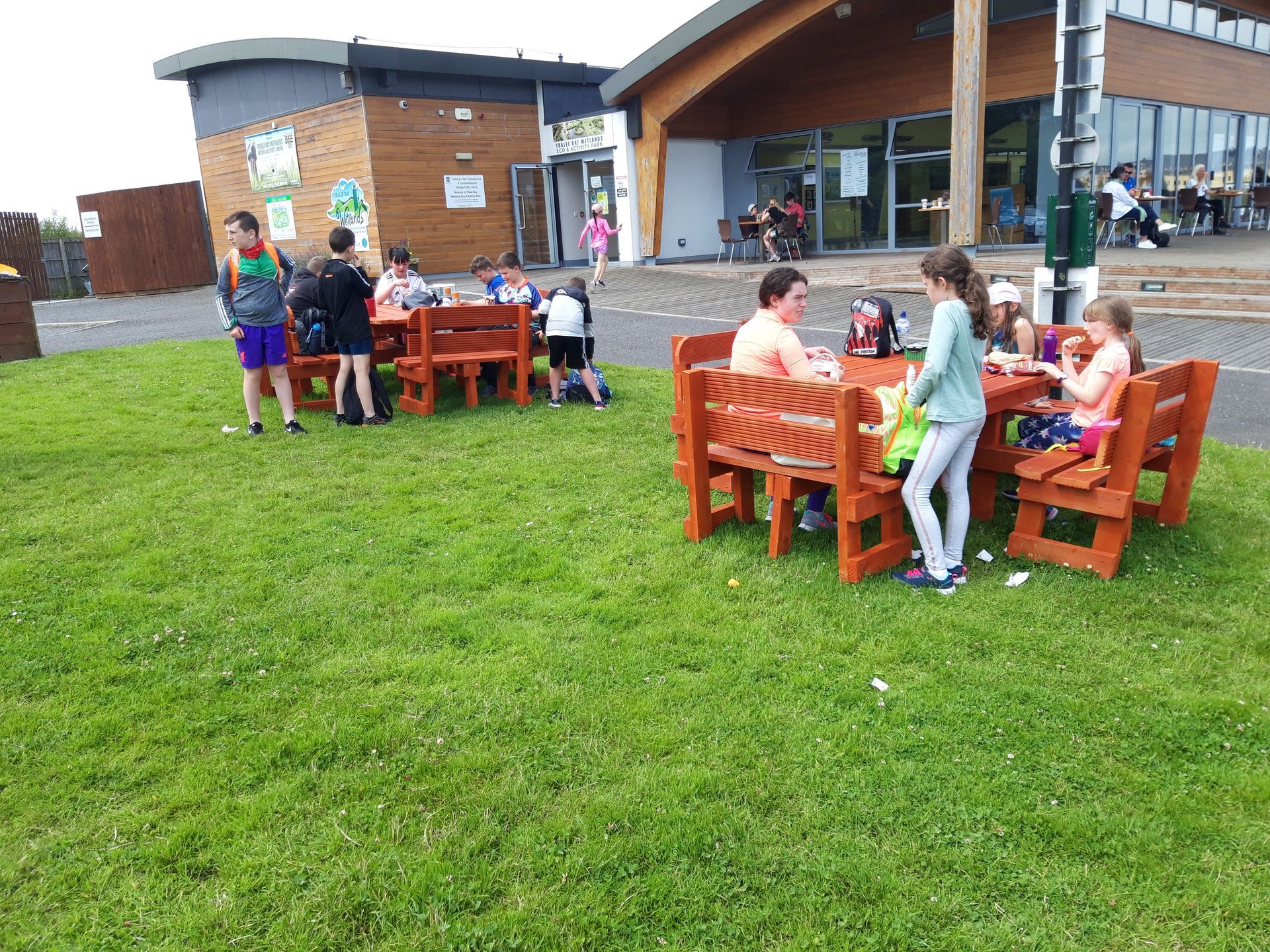 Children eating at wooden picnic tables on a grassy lawn outside a building.
