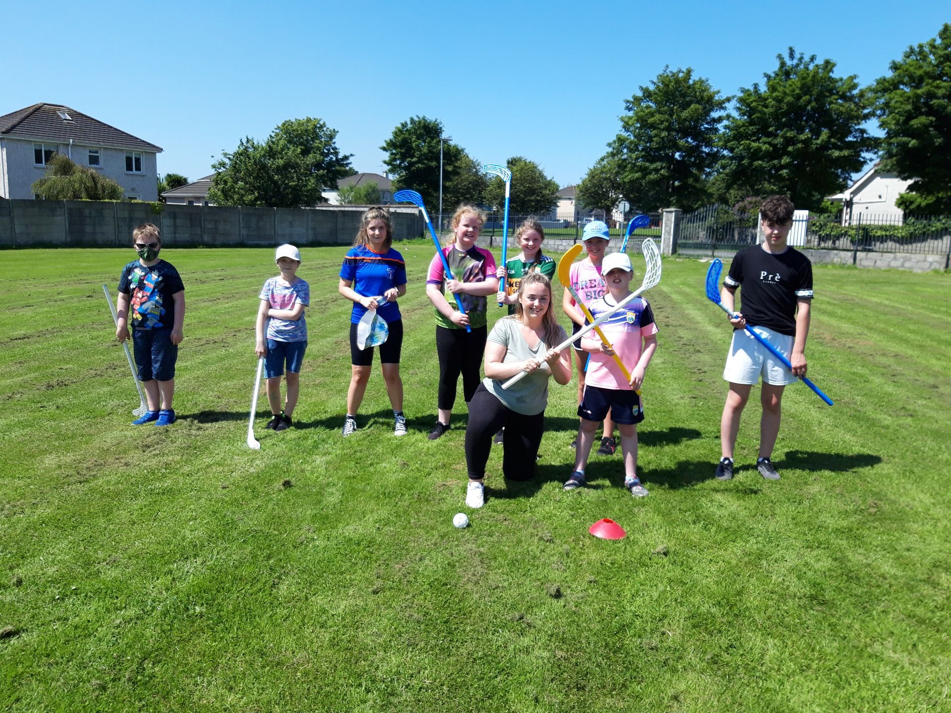 Group of kids on a green field holding sports sticks; one kneels with a ball, a sunny day.