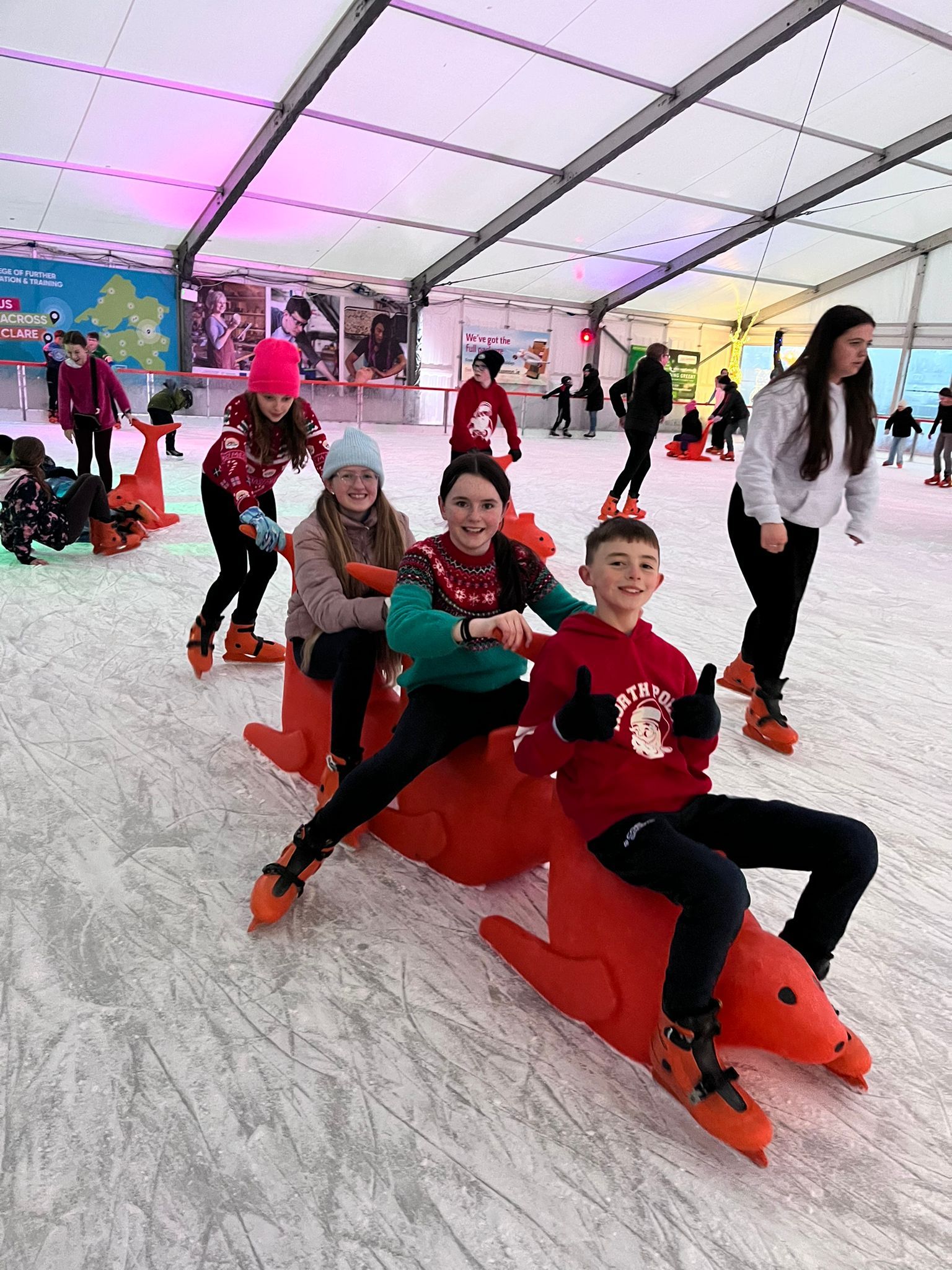 Children ice skating, laughing while sitting on red seals at an ice rink.