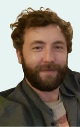 Man with curly brown hair and beard, smiling. Wearing a gray jacket over a patterned shirt.