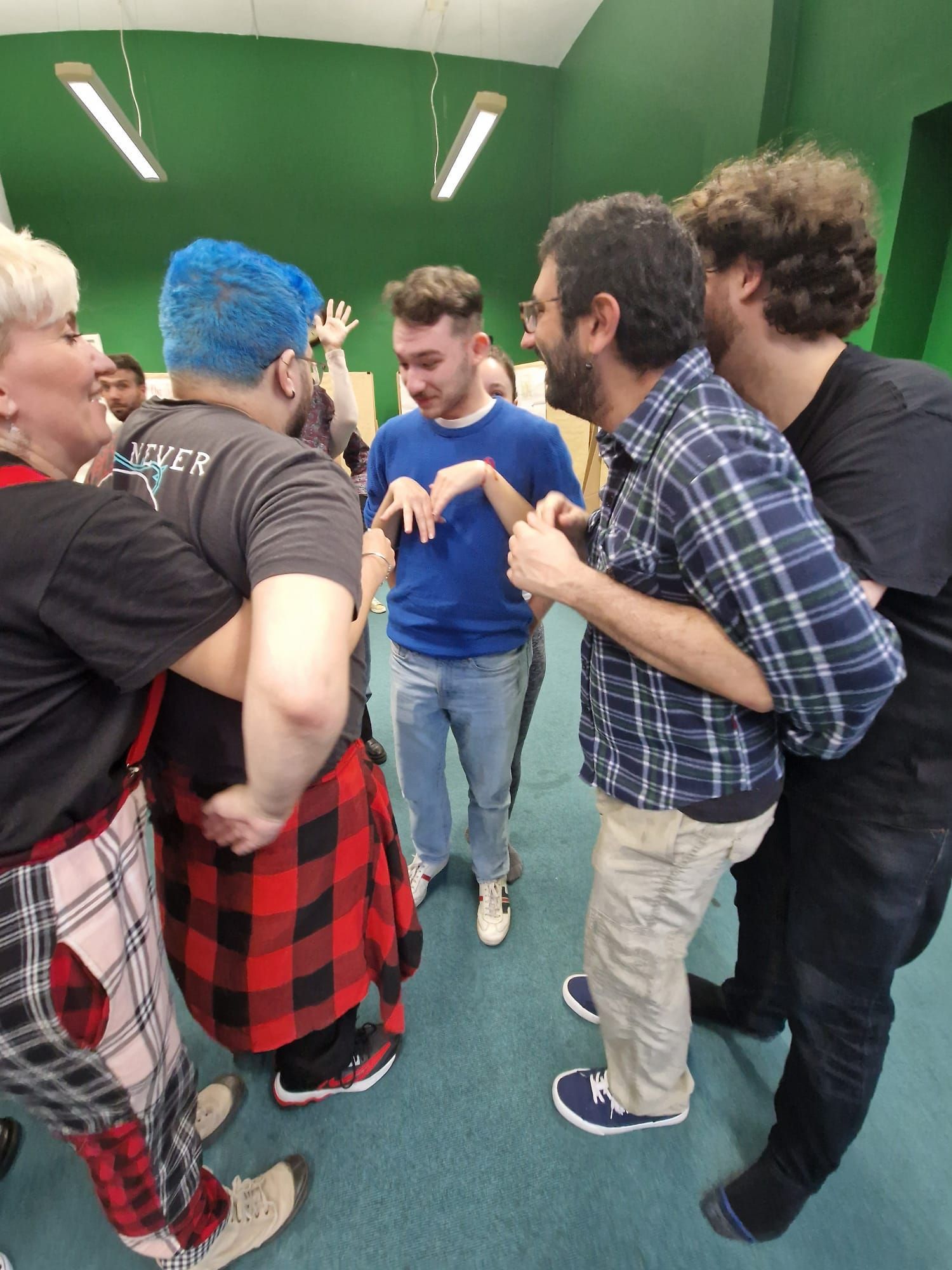 People in a circle, some hugging. A person with blue hair stands in the center, making a gesture. Bright room, green wall.