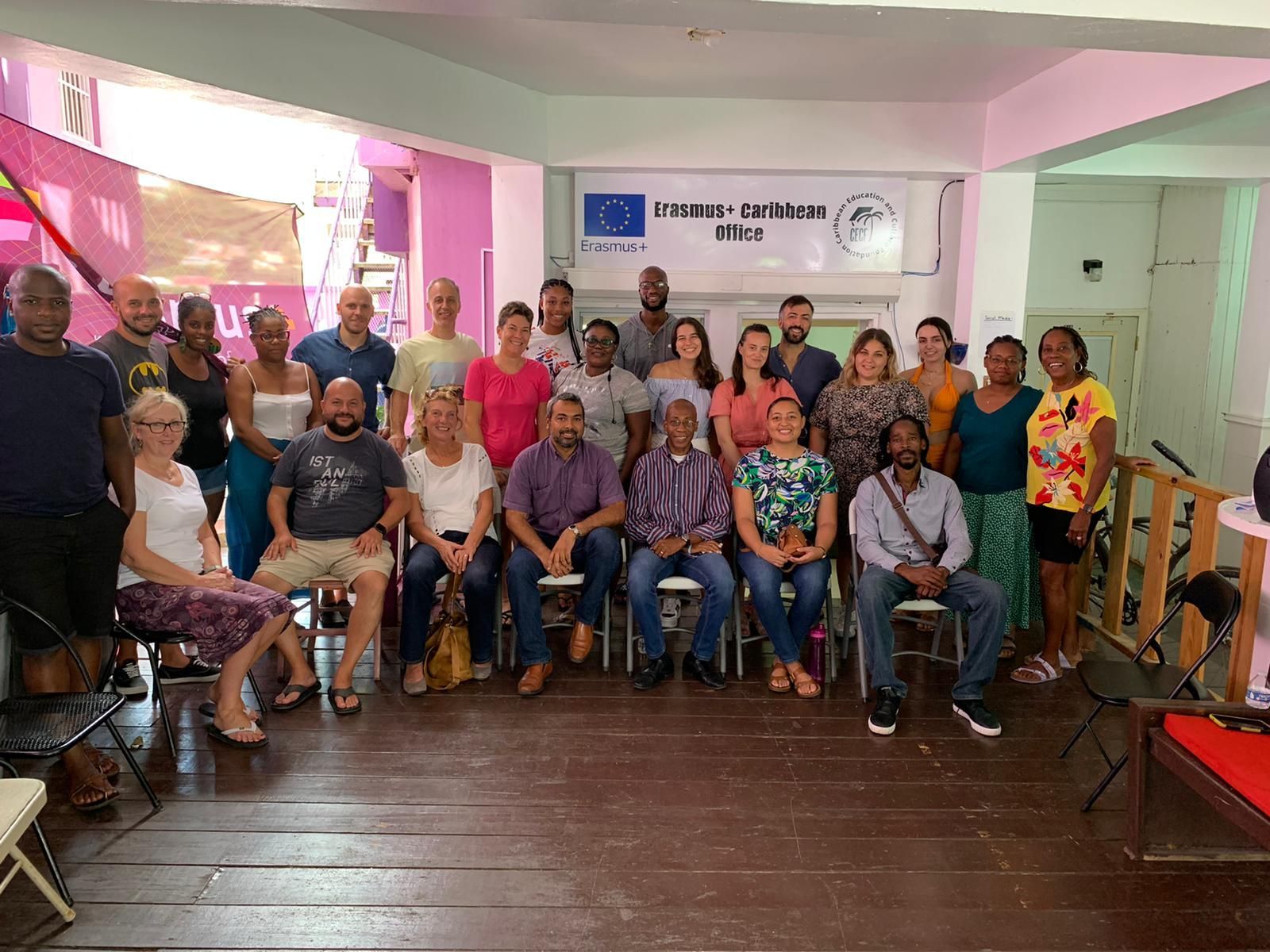 Group of people posing for a photo indoors, likely a meeting or workshop. White sign on the wall, wood floor.